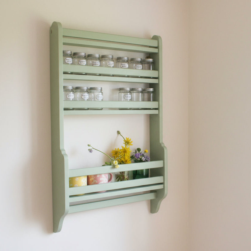 A green wooden  ladder-style tea-towel rail under a hanfdade, hand-painted spice rack, mounted on a white wall, with spice jar on the shelf section, mugs and a vase of yellow flowers.