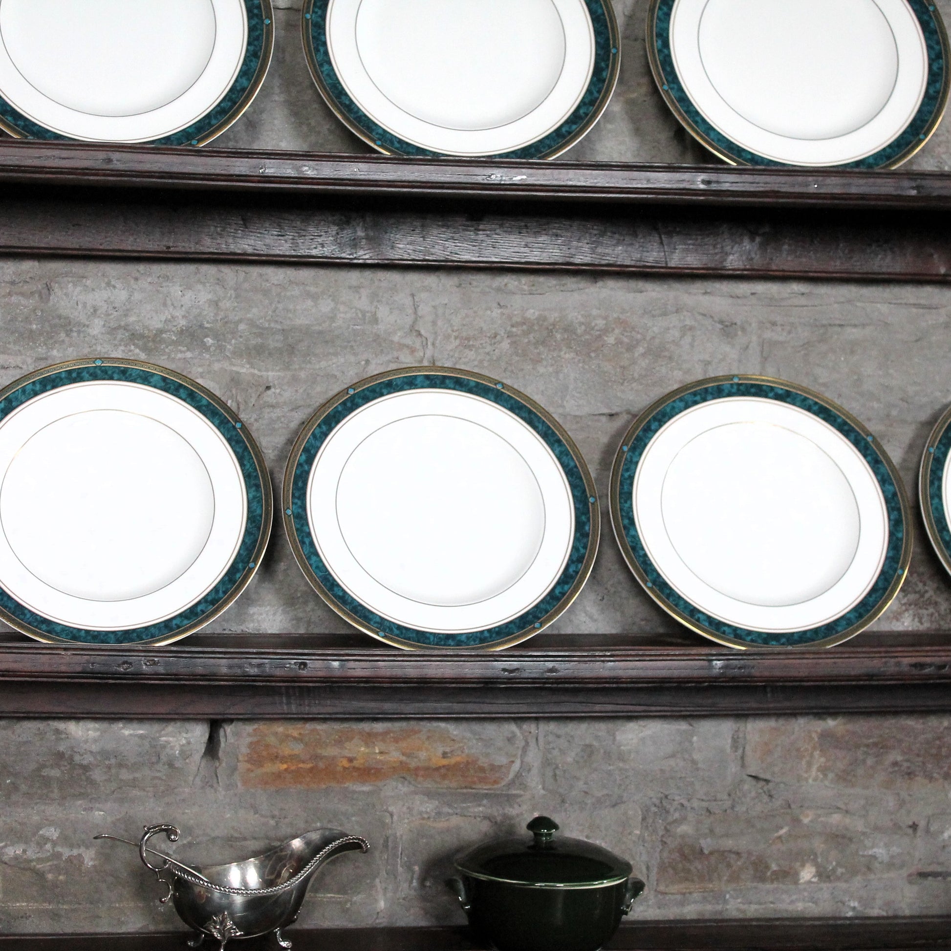 Set of white china plates with green rims on a metal shelf against a rustic stone wall.