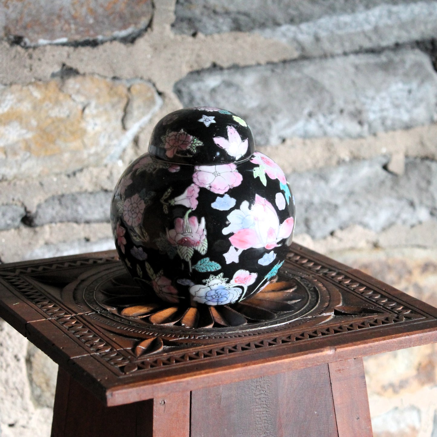 Chinese porcelain black ginger jar with flowers on a wooden pedestal against a stone wall
