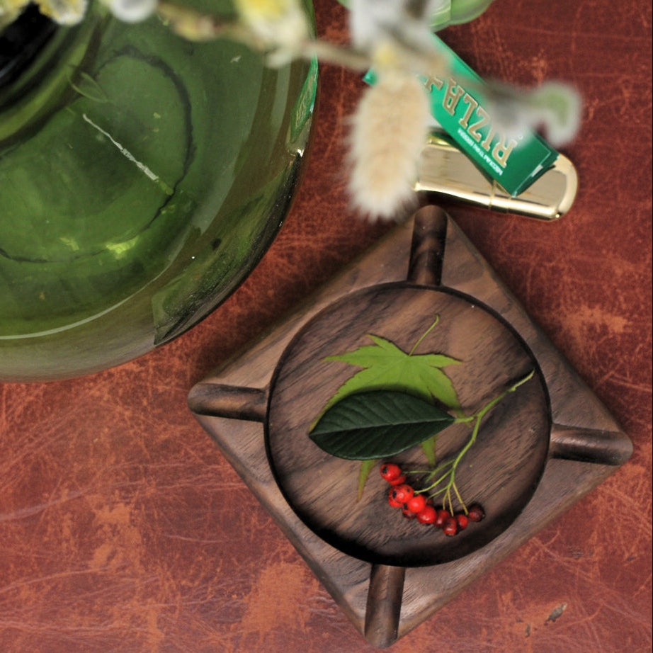 A square dark wood ashtray with a circular center insert, styled on a white surface with a green glass vase, dried grass, and a green lighter in the background.