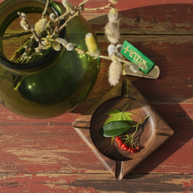 A square dark brown wooden ashtray with a circular center, styled with a green leaf and small red berries, shown next to a green glass vase with willow branches on a red surface.