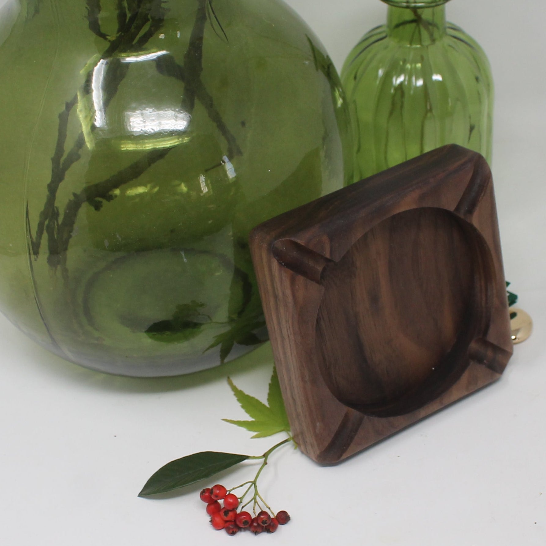 A green glass vase and a small green glass bottle with dried stems, alongside a dark brown wooden square ashtray, arranged on a white surface.