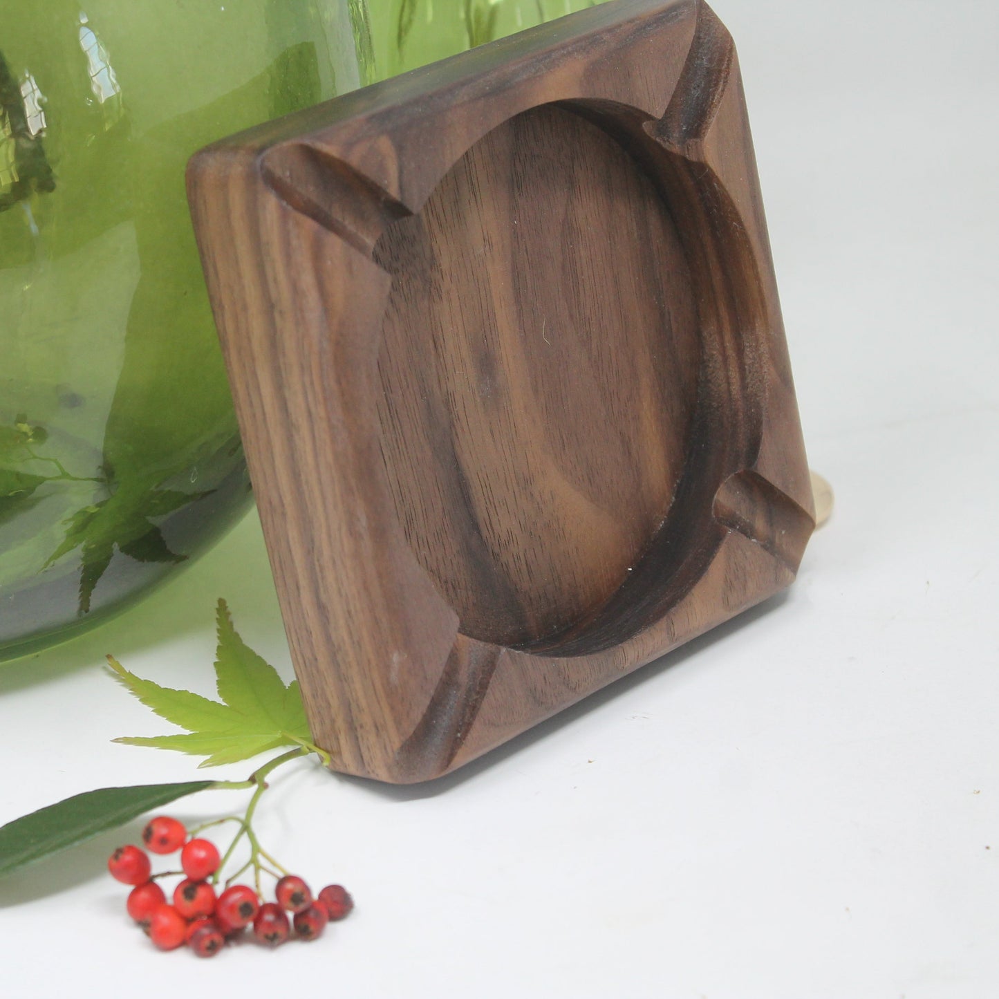 A square dark brown wooden ashtray with a circular center recess, shown on a white surface next to a green glass vase and small red berries.