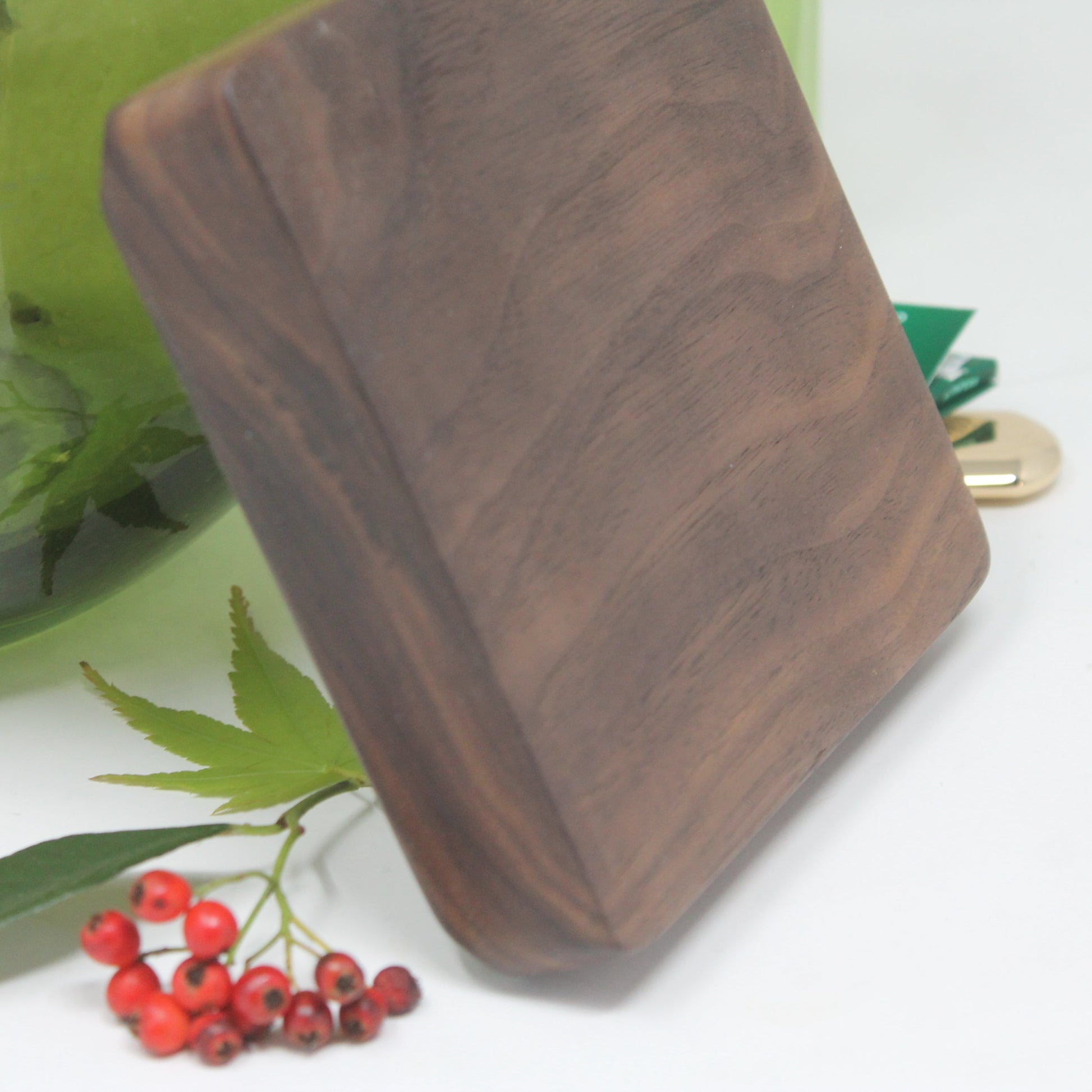 A square dark brown wooden cutting board with rounded corners, shown standing on a white surface next to green leaves and red berries, with a green glass container in the background.