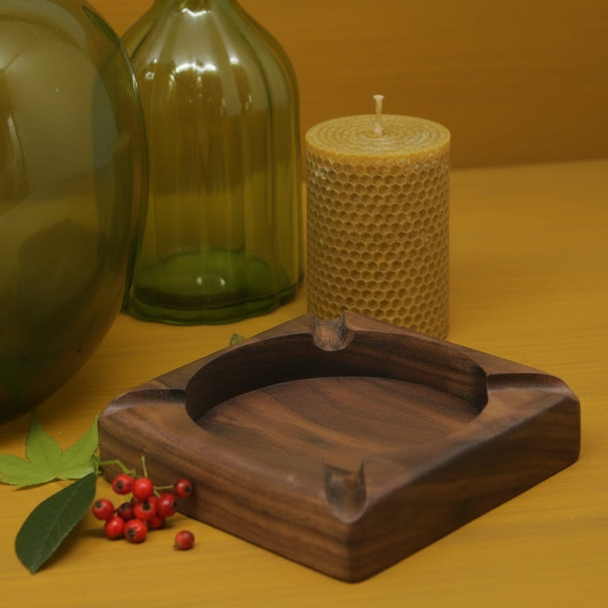 A square dark brown wooden ashtray with a polished finish and integrated ash bins, styled on a white surface with green glass bottles, red berries, and a green rolling paper pack in the background.