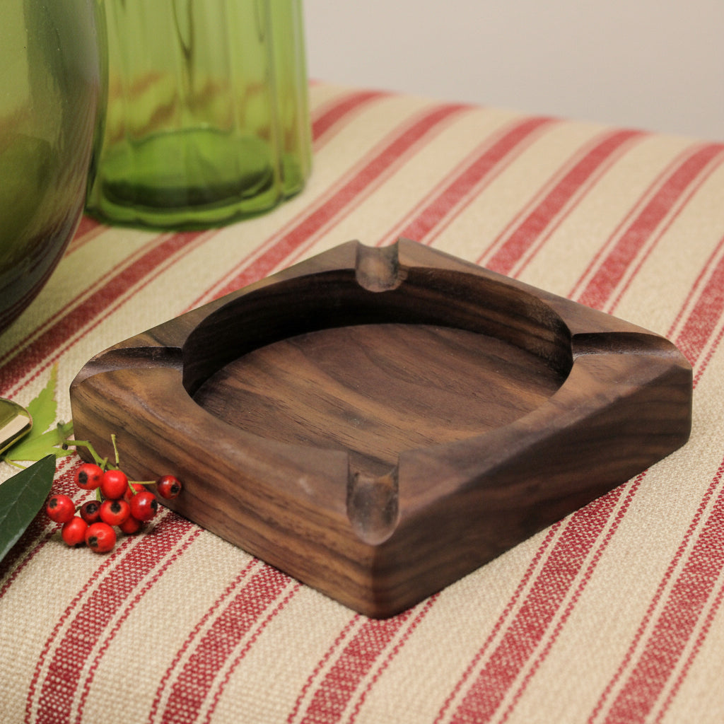 A square dark brown wooden ashtray with a carved circular center recess and four angled smoking slots, shown on a white surface with decorative green glass bottles and red berries in the background.