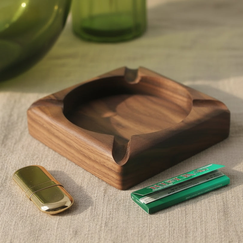 A square dark brown wooden ashtray with integrated ash pockets, styled on a white surface with a green glass vase, red berries, and two small metallic cases.