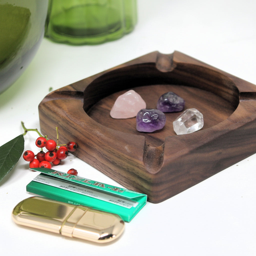 A square dark wood ashtray with a carved circular center recess, styled on a white surface with a gold lighter, a green cigarette pack, and red berries nearby.