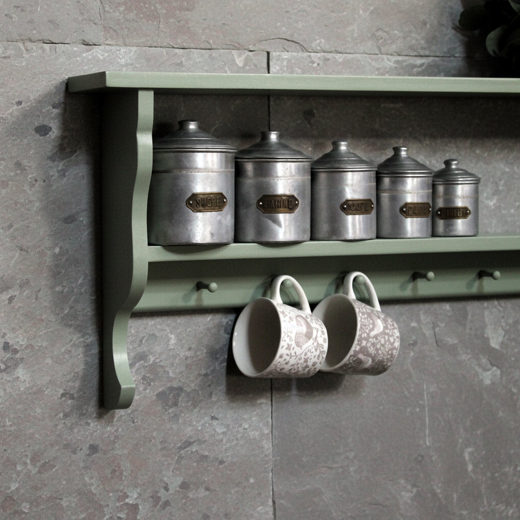 A sage green wooden wall-mounted shelf with two tiers, holding five metallic canisters and two patterned ceramic mugs hanging from side hooks against a white wall.