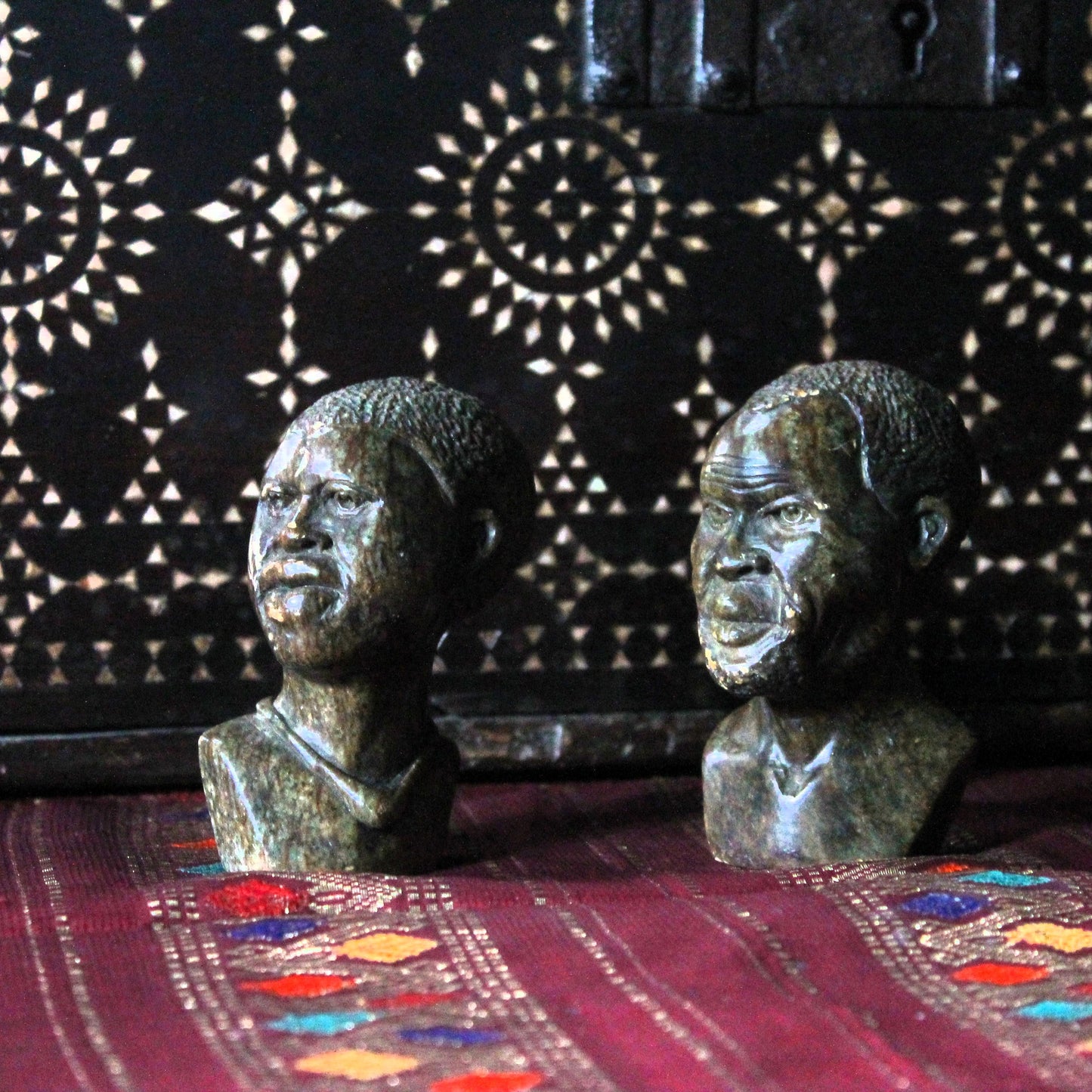 Two Kenyan kisii stone carvings  busts on a textured surface with a patterned background