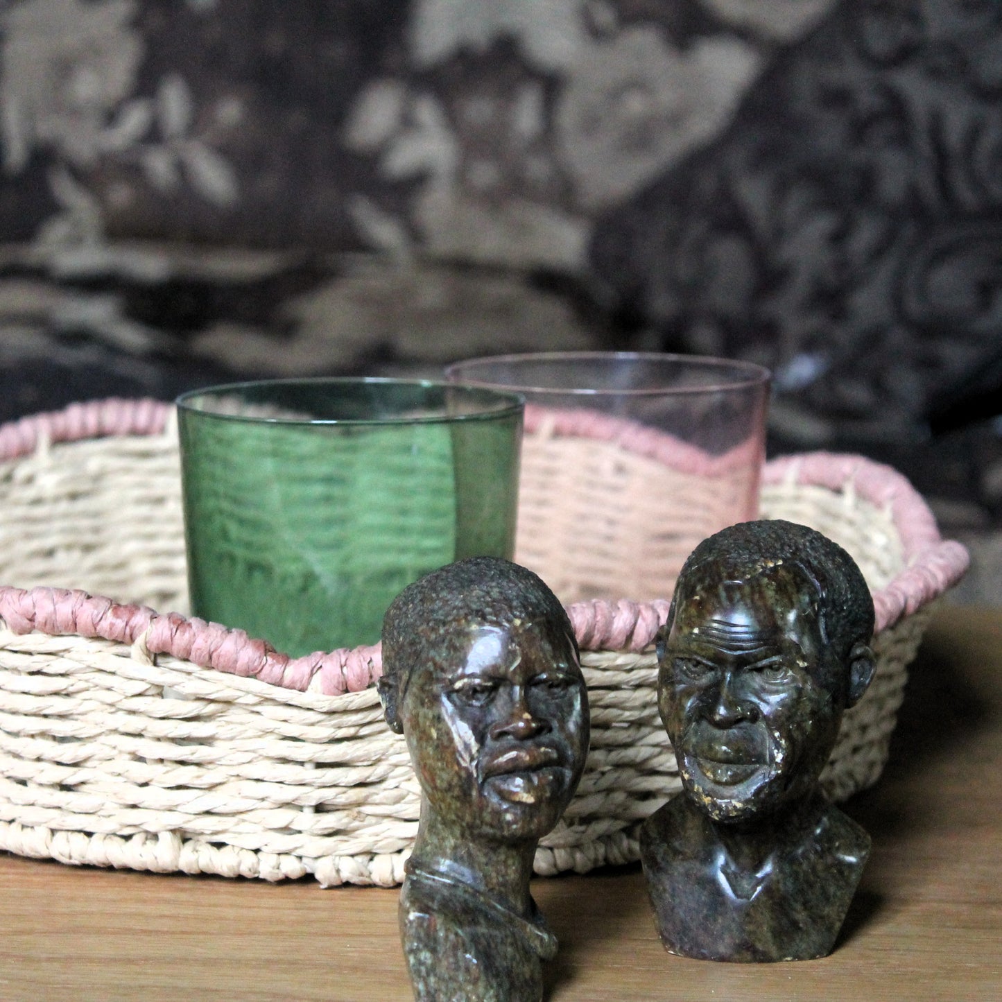 Two vintage African art  head sculptures in front of a woven basket with green and pink glasses. 