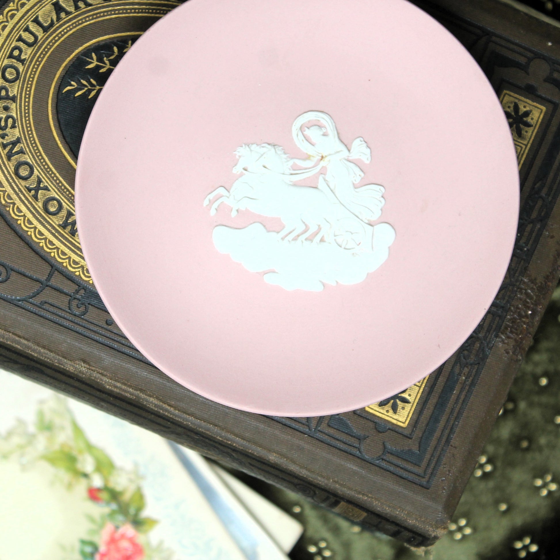 A round pink Wedgwood ring dish or photographed on a dark patterned book.