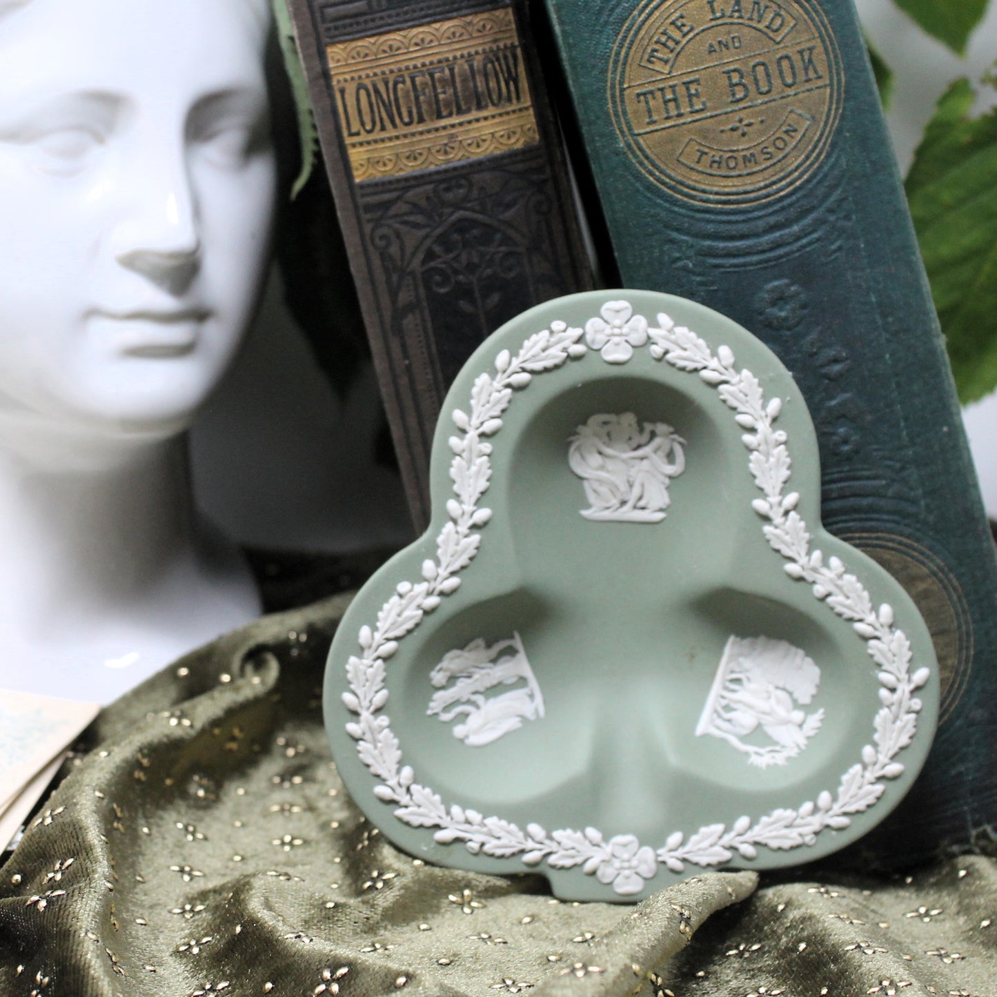 Decorative green and white ceramic dish with classical design on a textured surface with books and statue in the background.