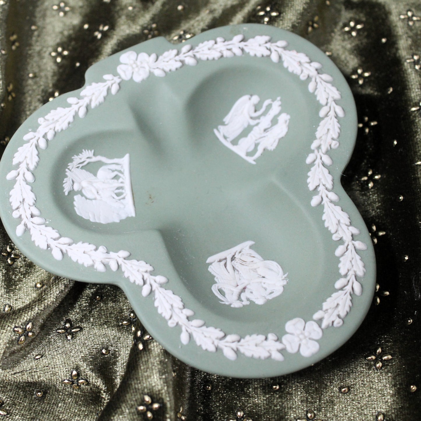 Decorative green Wedgewood trinket dish with white floral and leaf patterns on a textured green fabric background