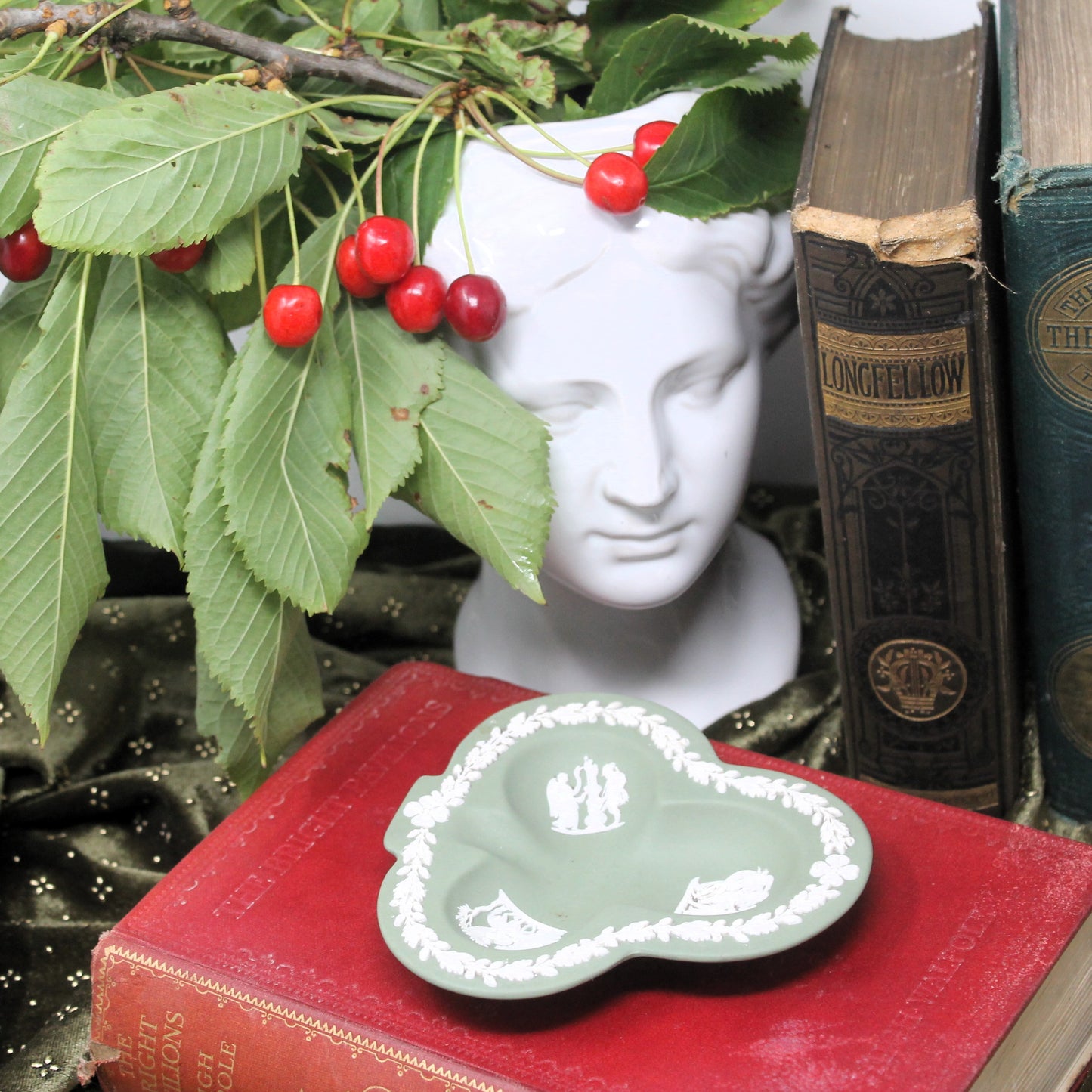 Decorative scene with cherries, a bust, books, and a green Wedgewood  jasperware trefoil dish on a red book.