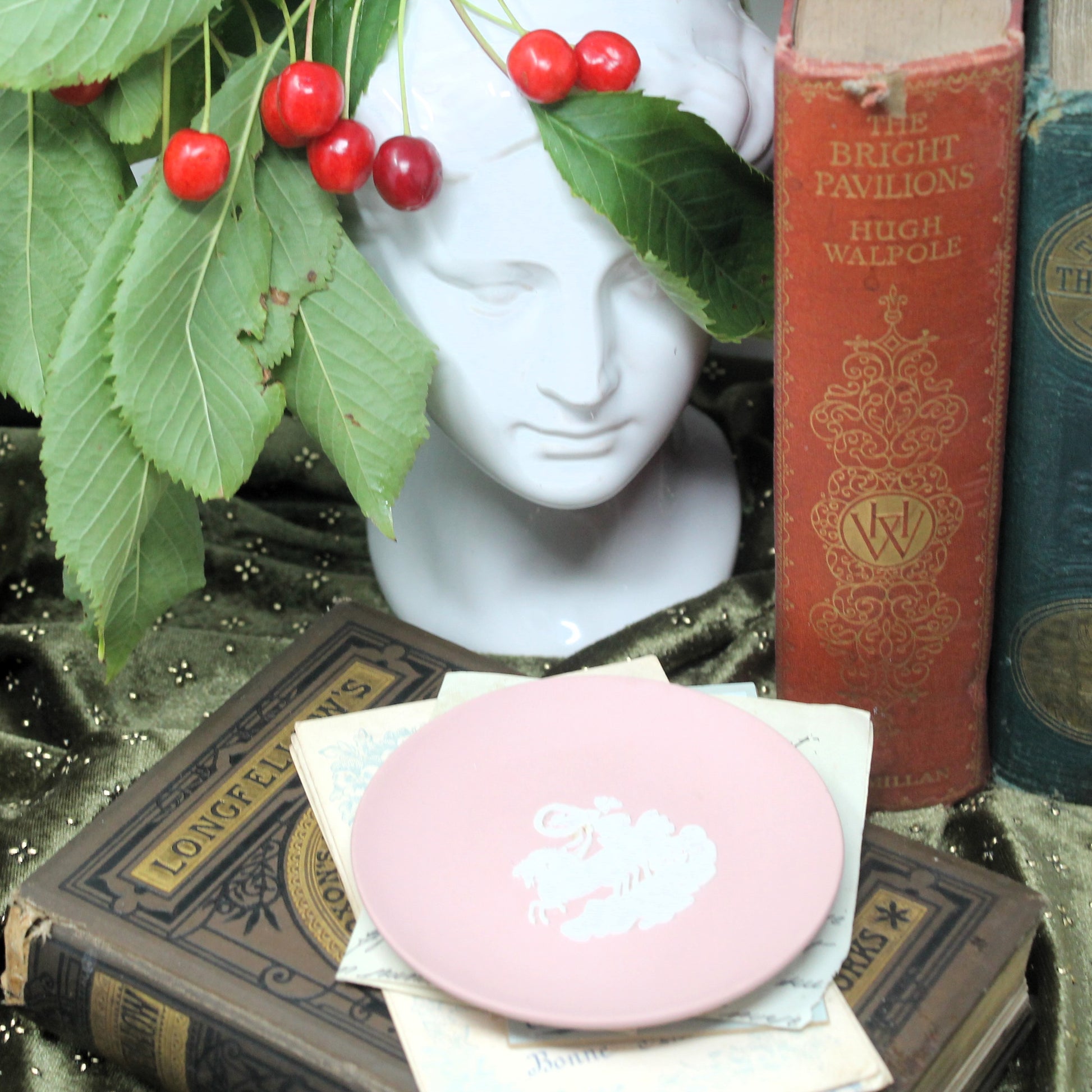 A round light pink Wedgwood ring dish  with a white charioter and horses pattern, styled on vintage books next to a white bust sculpture with red cherries and an ornate red book.