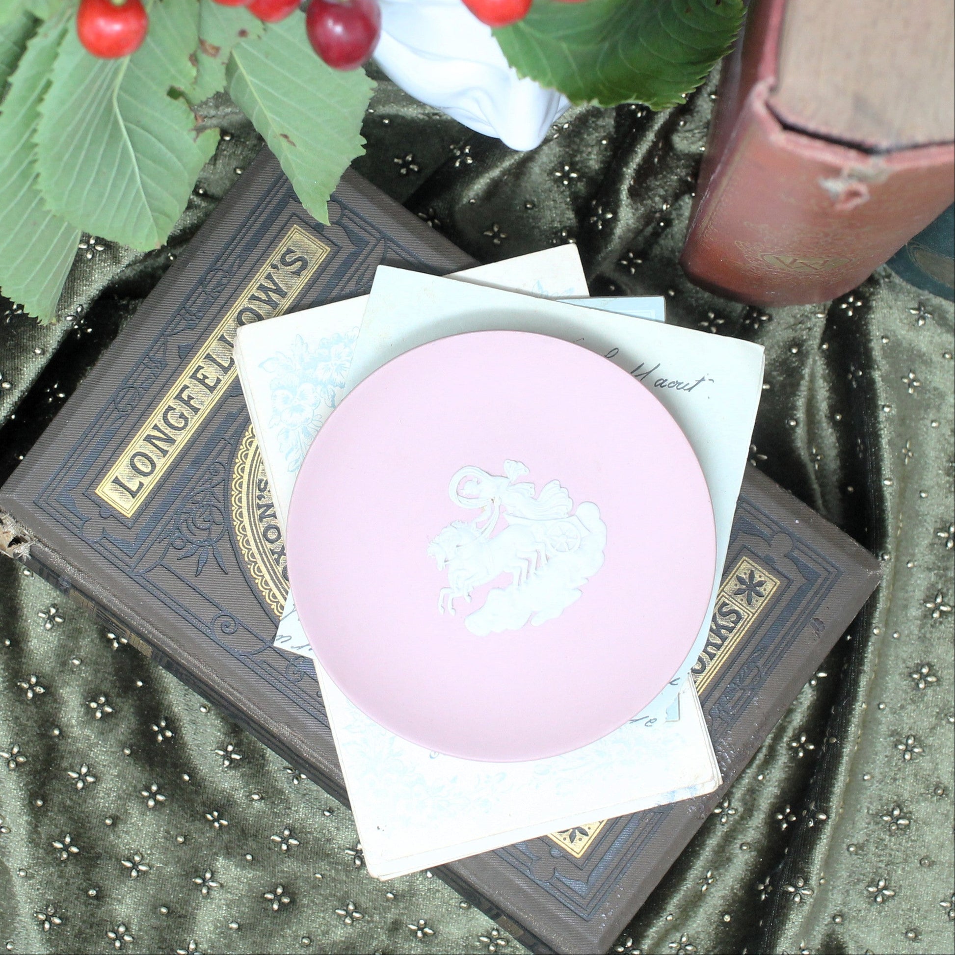 A round dusky pink small Wedgwood jasperware dish   with a white embossed crest, resting on white tissue paper atop a dark brown vintage book. The scene is styled with green leaves and red cherries on a dark green patterned fabric background.