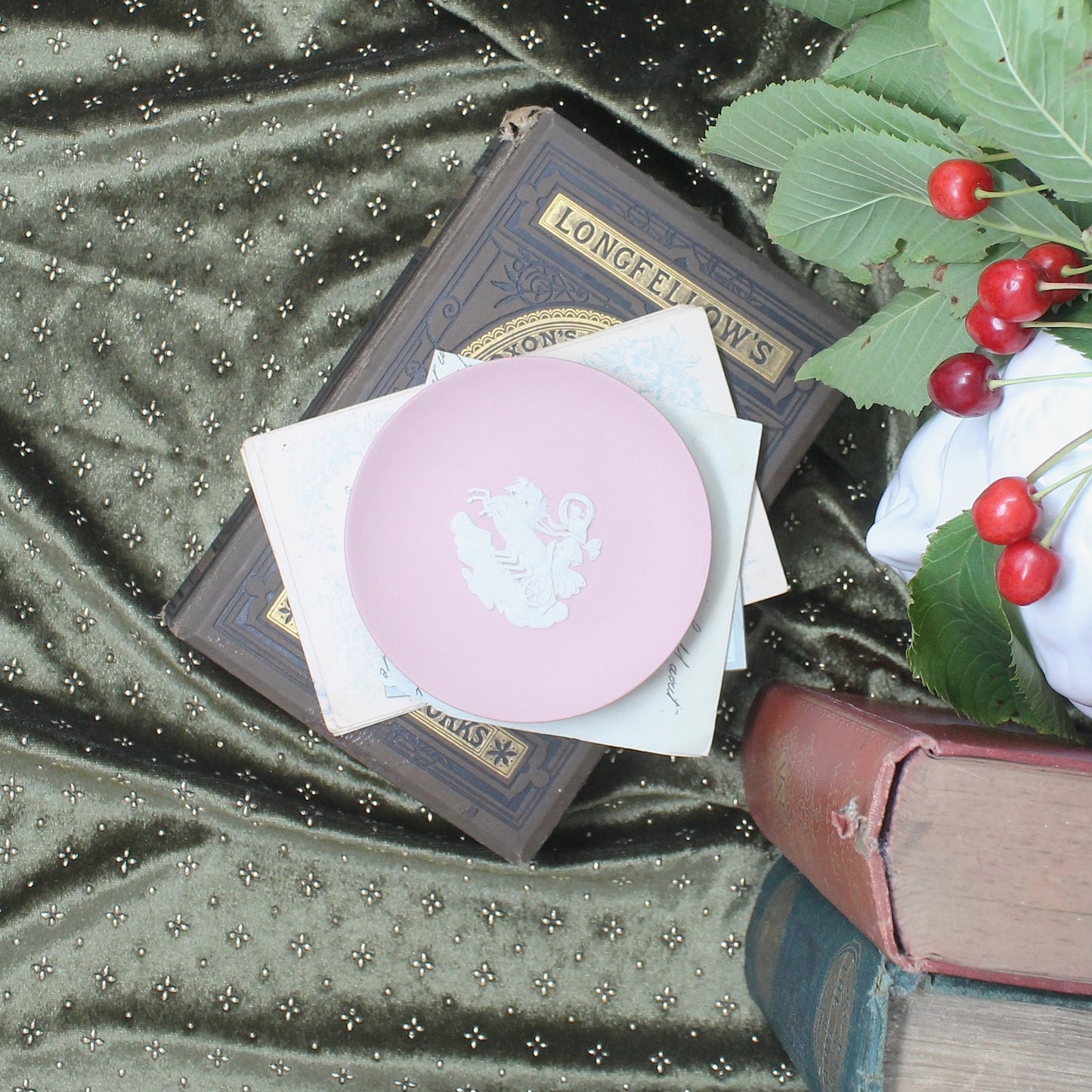 A round pink wedgwood keepsake dish with a white applied scene, resting on vintage books and white envelopes over a green patterned fabric, with red cherries and white flowers at the side.