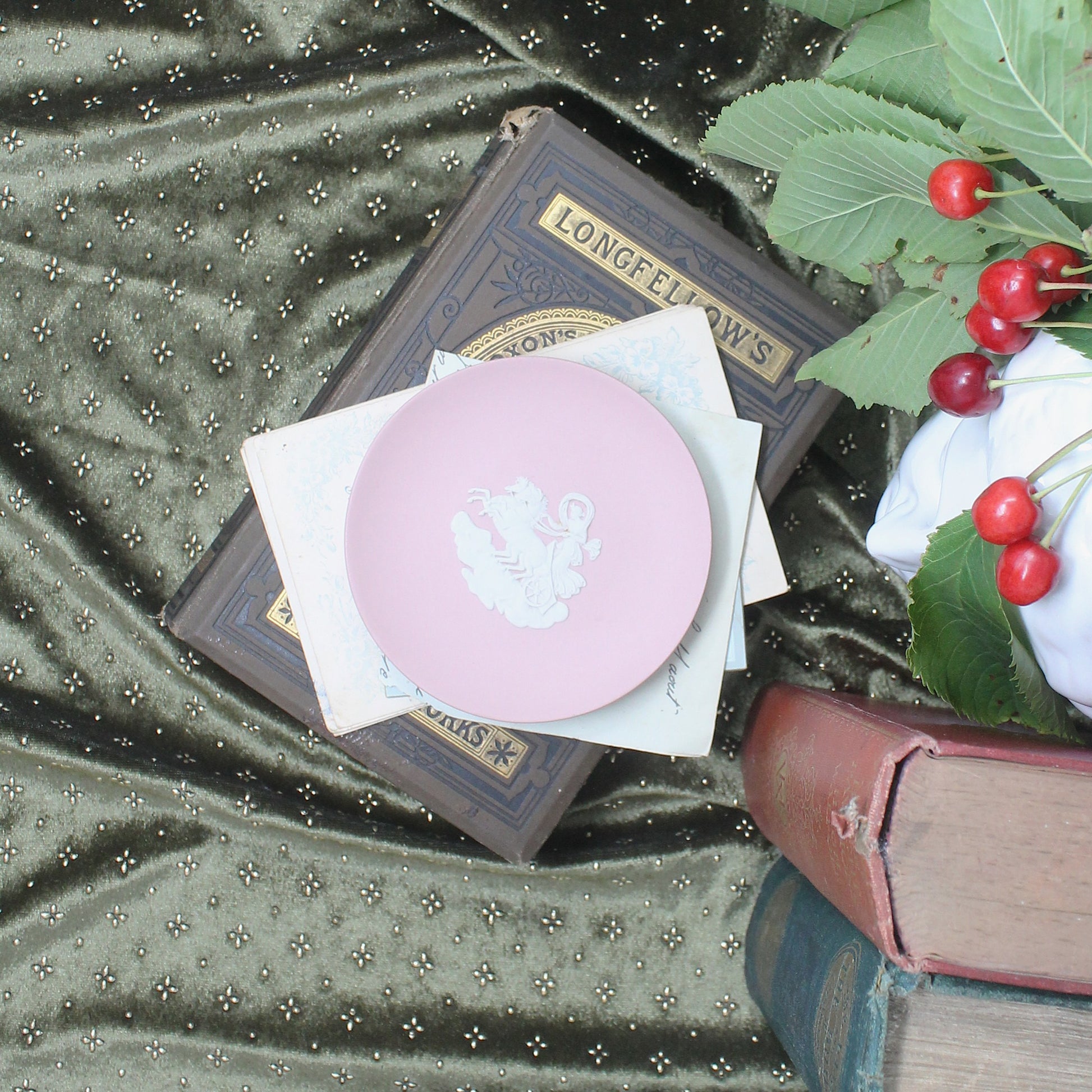 A round pink wedgwood keepsake dish with a white applied scene, resting on vintage books and white envelopes over a green patterned fabric, with red cherries and white flowers at the side.