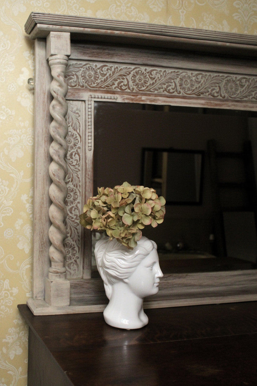 Decorative mirror with intricate carvings on a wall, featuring a white vase with flowers on a dark surface.