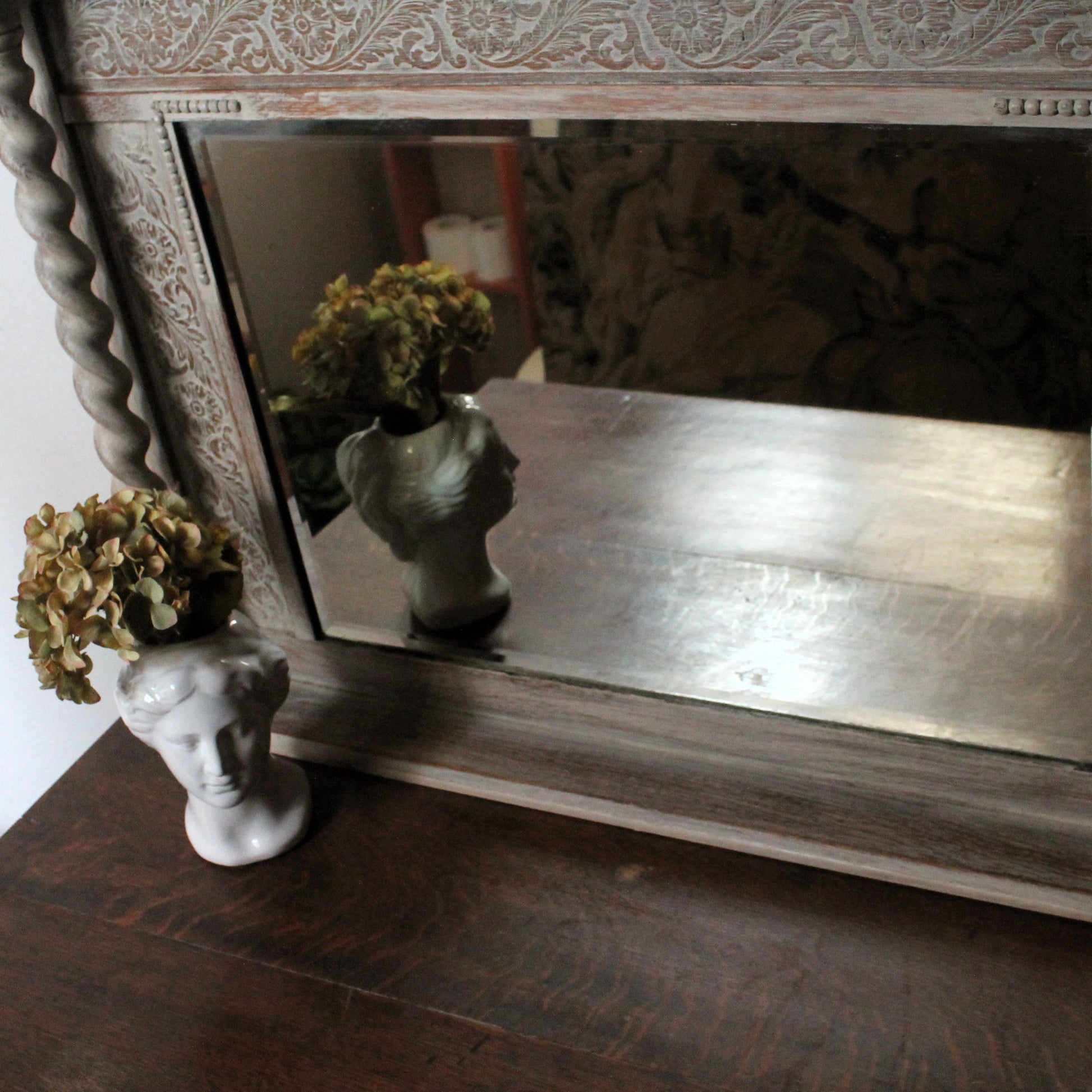 Decorative mirror with a wooden frame on a surface, featuring a small bust and flowers.