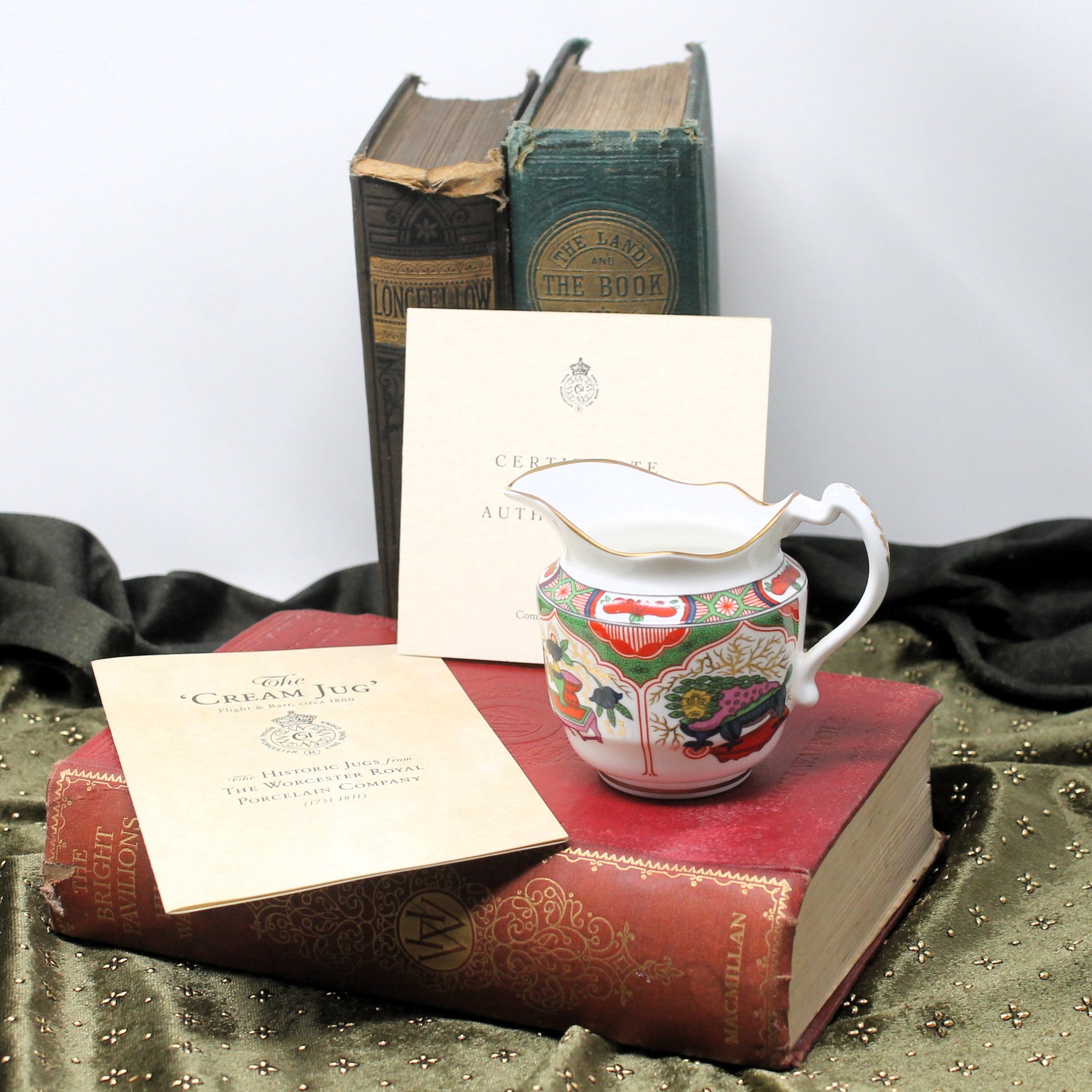 A decorative porcelain creamer pitcher with a multicolor floral and bird pattern, displayed on top of a red vintage book. In front is a cream-colored certificate card, and behind it are two antique books, one dark brown and one green.