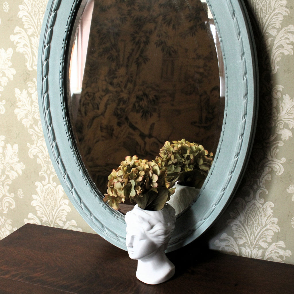 Round mirror with decorative frame on a wall, reflecting a patterned curtain.
