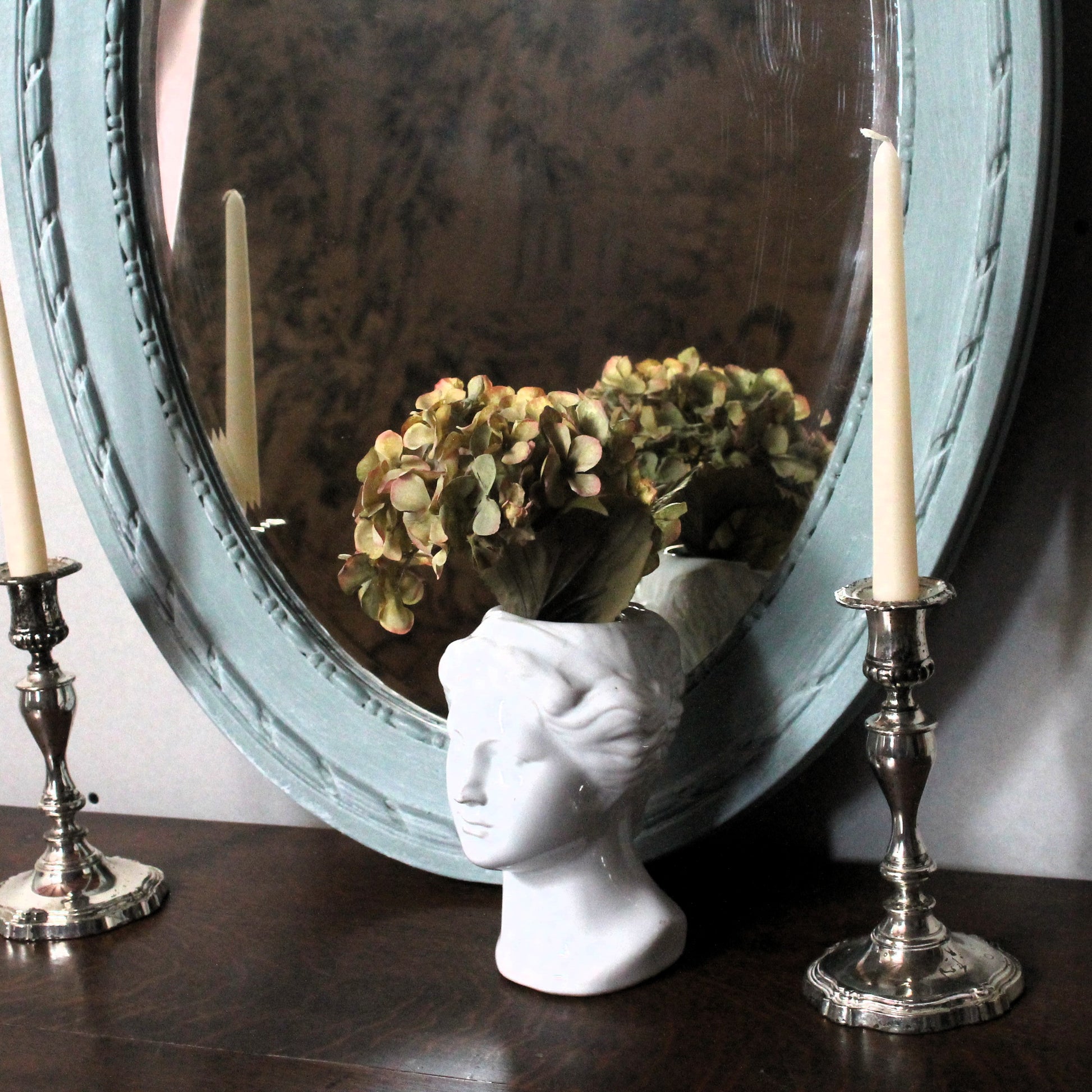 Decorative setup with a mirror, candles, and a vase shaped like a face on a wooden surface.