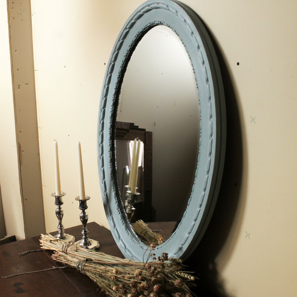 Round mirror with a light blue frame on a wall, with candles and a broom in the foreground.