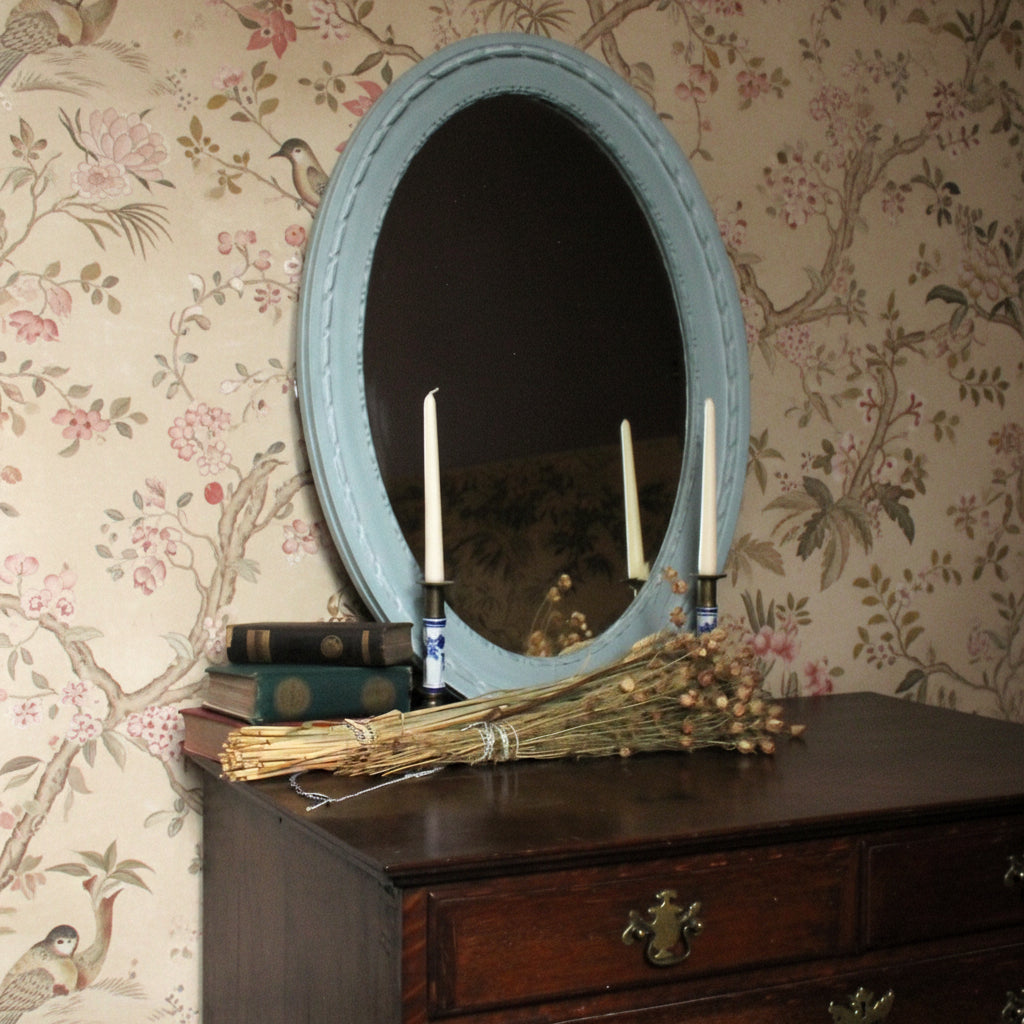 Oval mirror with decorative elements on a wooden surface against a plain wall.