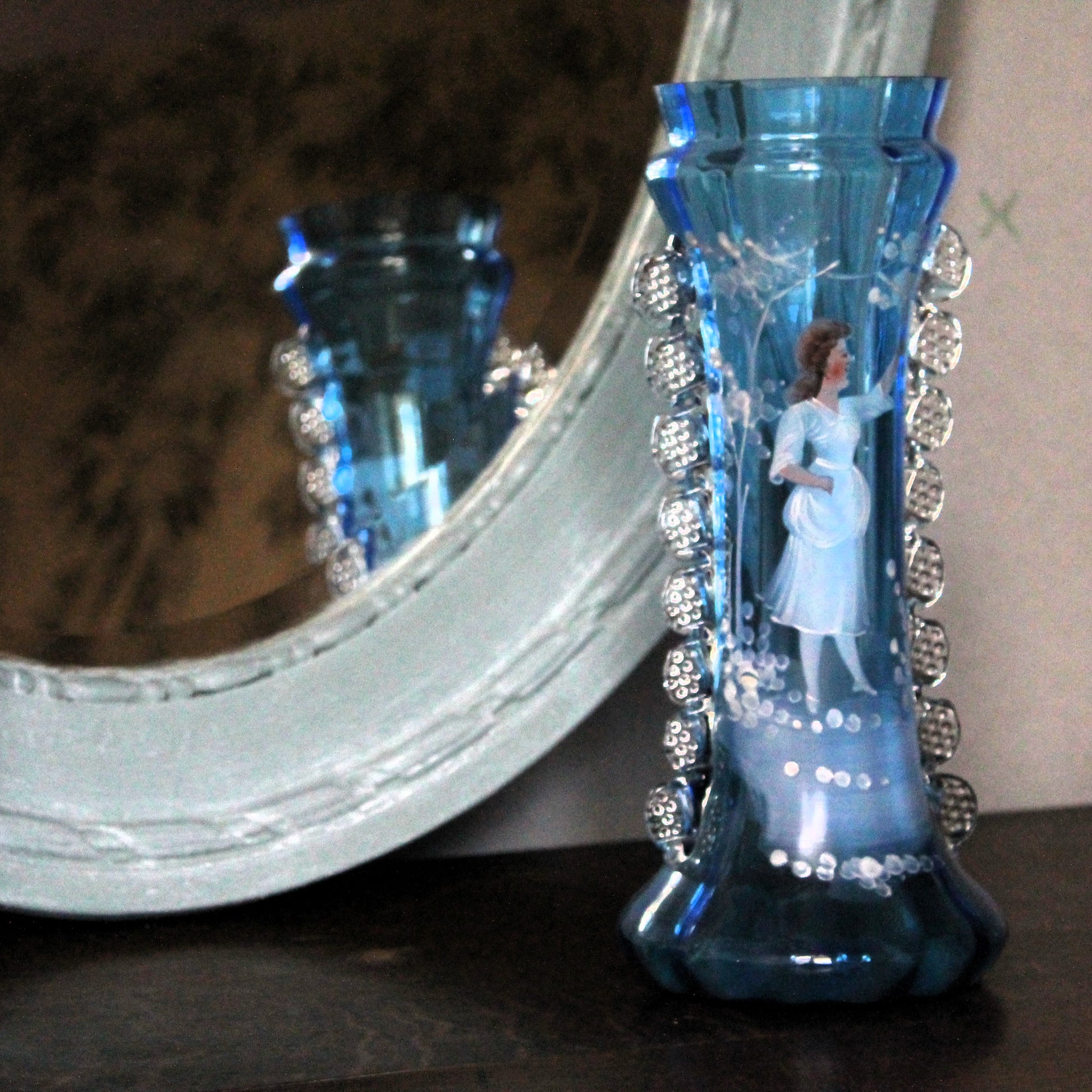 A decorative blue glass vase with a printed fairy figure inside and silver beaded trim, displayed next to a large ornate mirror on a dark surface.