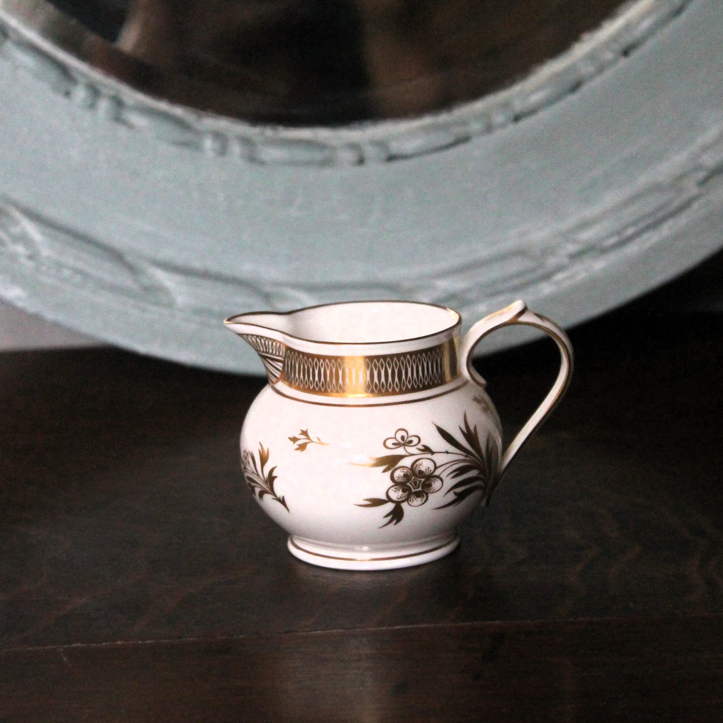 A small white porcelain creamer pitcher with gold trim and floral decoration, set on a dark wooden surface with a large light blue mirror in the background. Royal Worcester Grainger jug