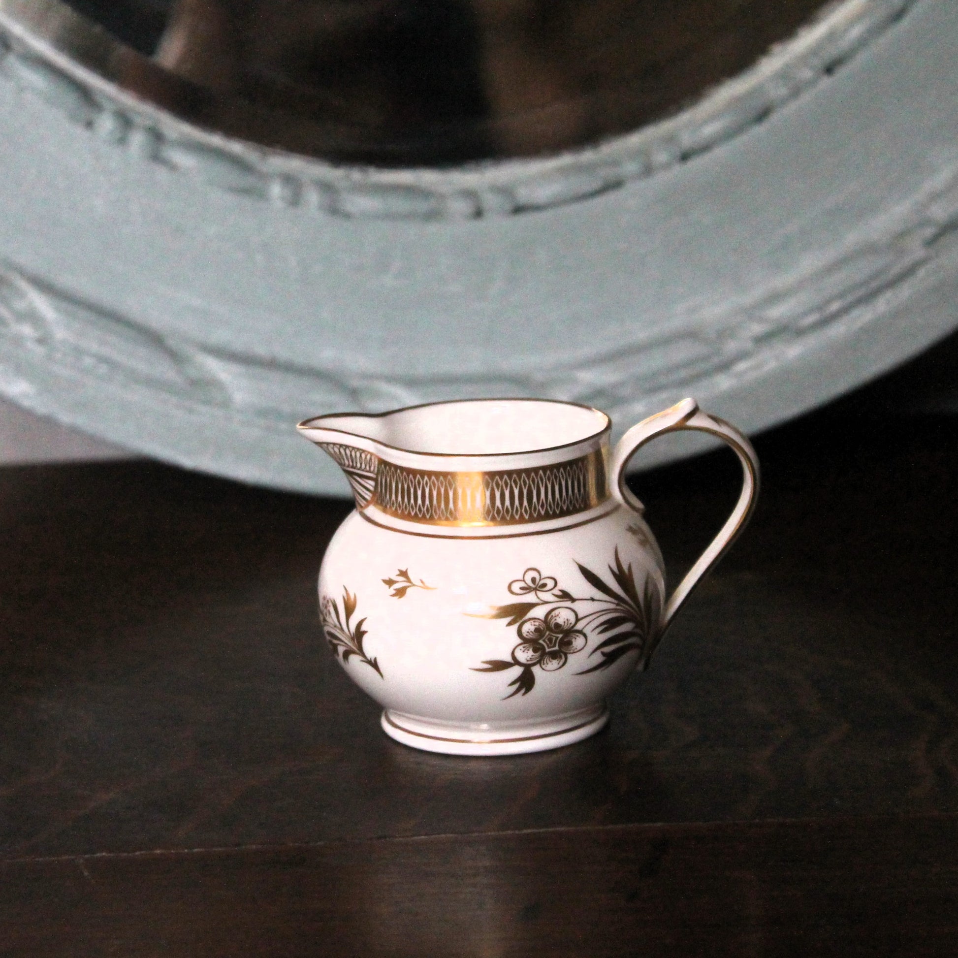 A small white porcelain creamer pitcher with gold trim and floral decoration, set on a dark wooden surface with a large light blue mirror in the background. Royal Worcester Grainger jug