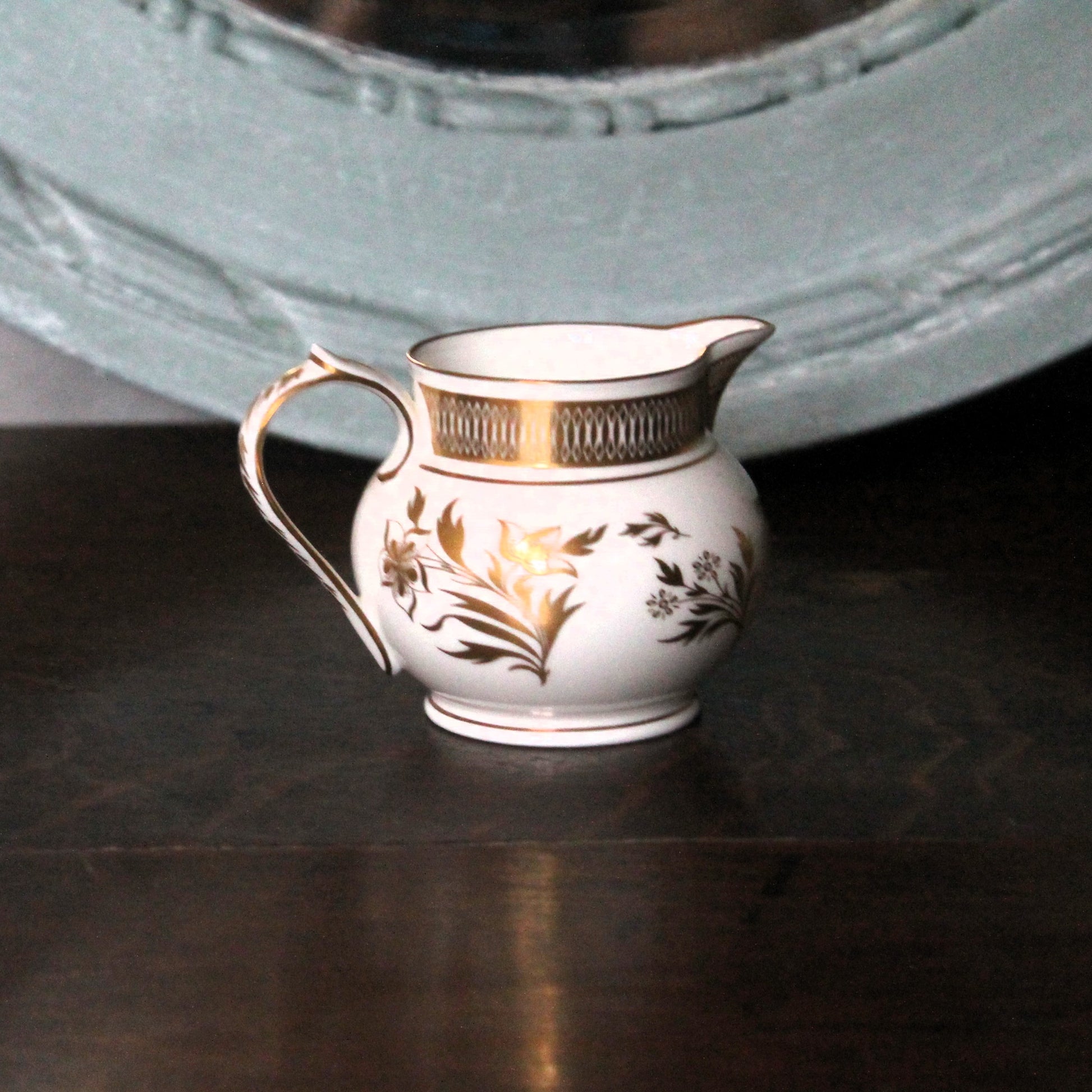 The Grainger jug, Royal Worcester  small white porcelain creamer jug with gold floral detailing and a gold band around the rim, resting on a dark wooden surface with a blurred teal mirror in the background.