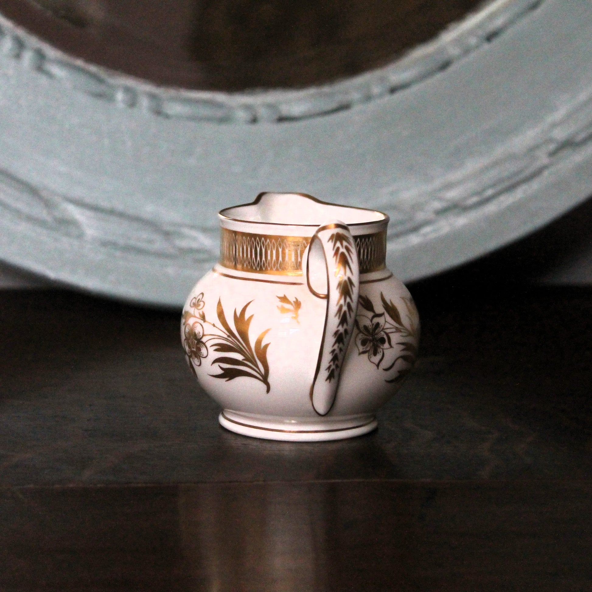 A small white porcelain creamer pitcher with gold and brown floral detailing, set on a dark reflective surface with a blurred light blue-gray decorative object in the background. Royal Worcester Grainger jug.