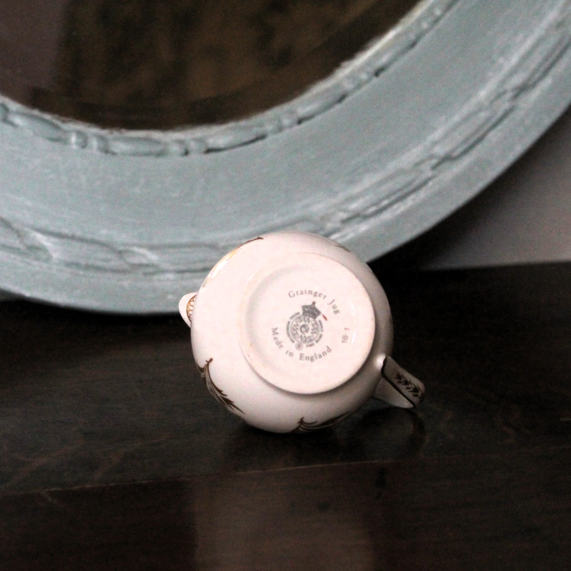 A small white porcelain teacup shown upside down on a dark wooden surface, with its printed bottom facing up. The cup has delicate gold floral detailing and a silver band near the rim.