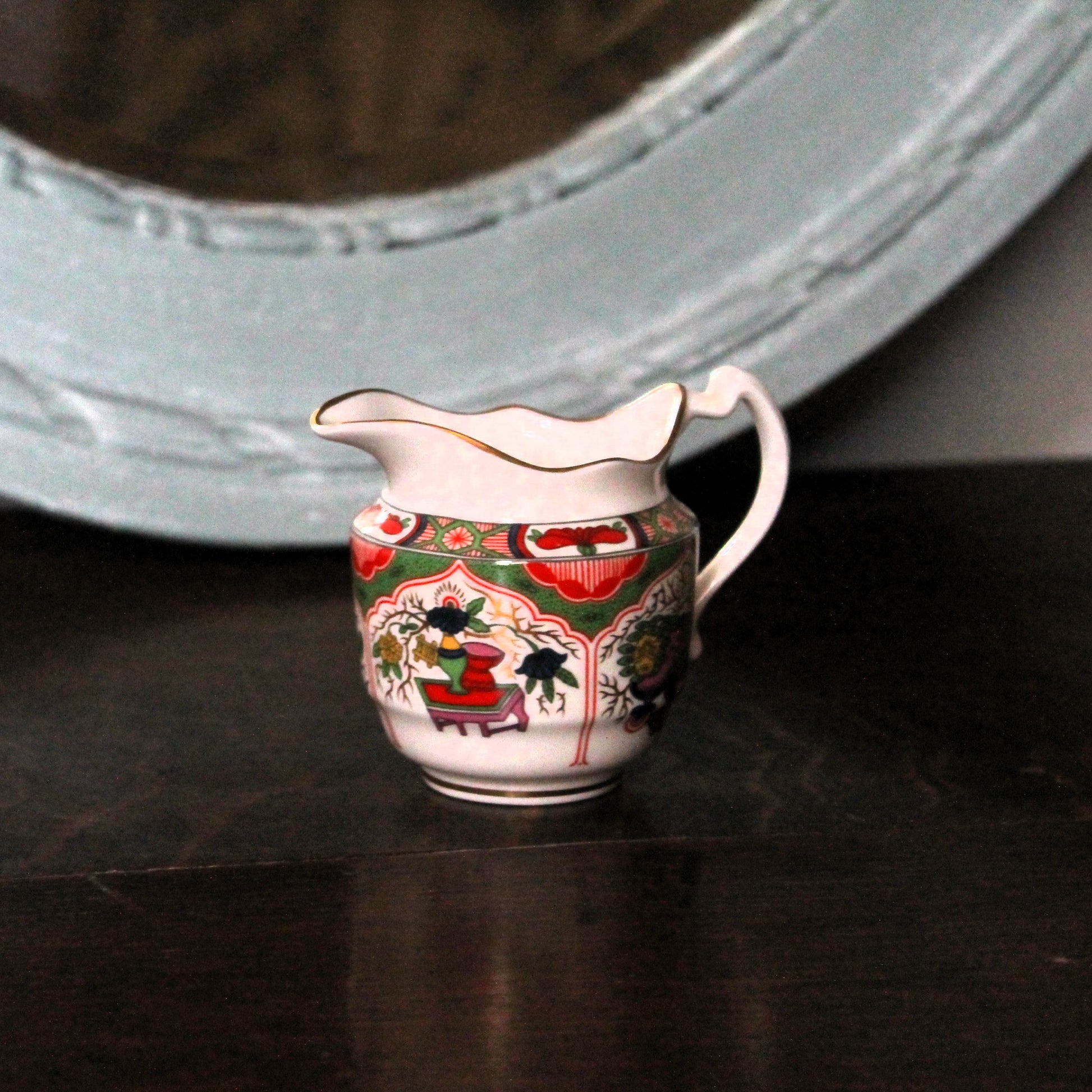 A small white Royal Worcester miniature porcelain creamer pitcher with a colorful hand-painted chinoiserie floral and figure pattern, resting on a dark wooden surface in front of a light blue mirror.