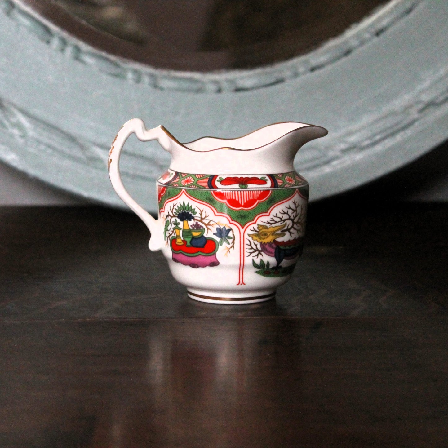 A small white porcelain creamer pitcher with a colorful hand-painted Dragons i  Compartments patterns of floral and landscape design, featuring red, green, yellow, and blue details, set on a dark wooden surface with a blurred light blue mirror in the background.