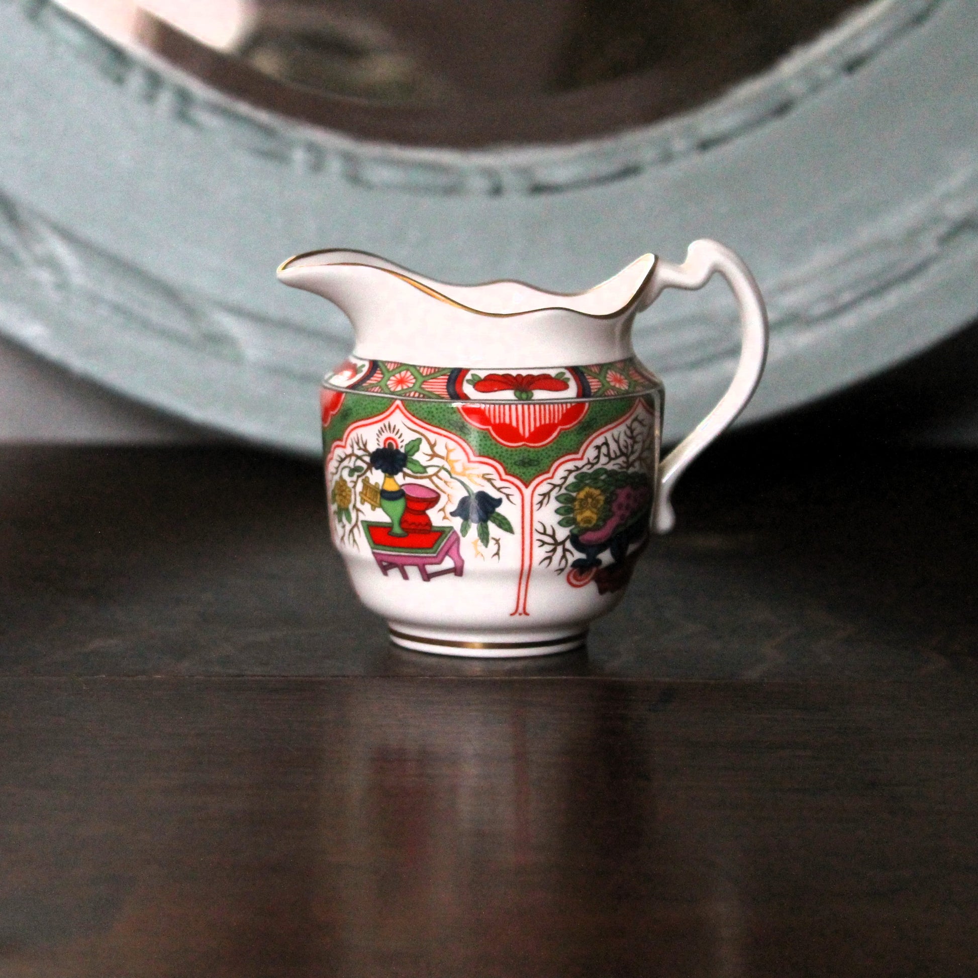 A small white chinese style Worcester porcelain creamer pitcher with a colorful floral and ornamental pattern, resting on a dark wooden surface with a blurred light blue decorative object in the background.