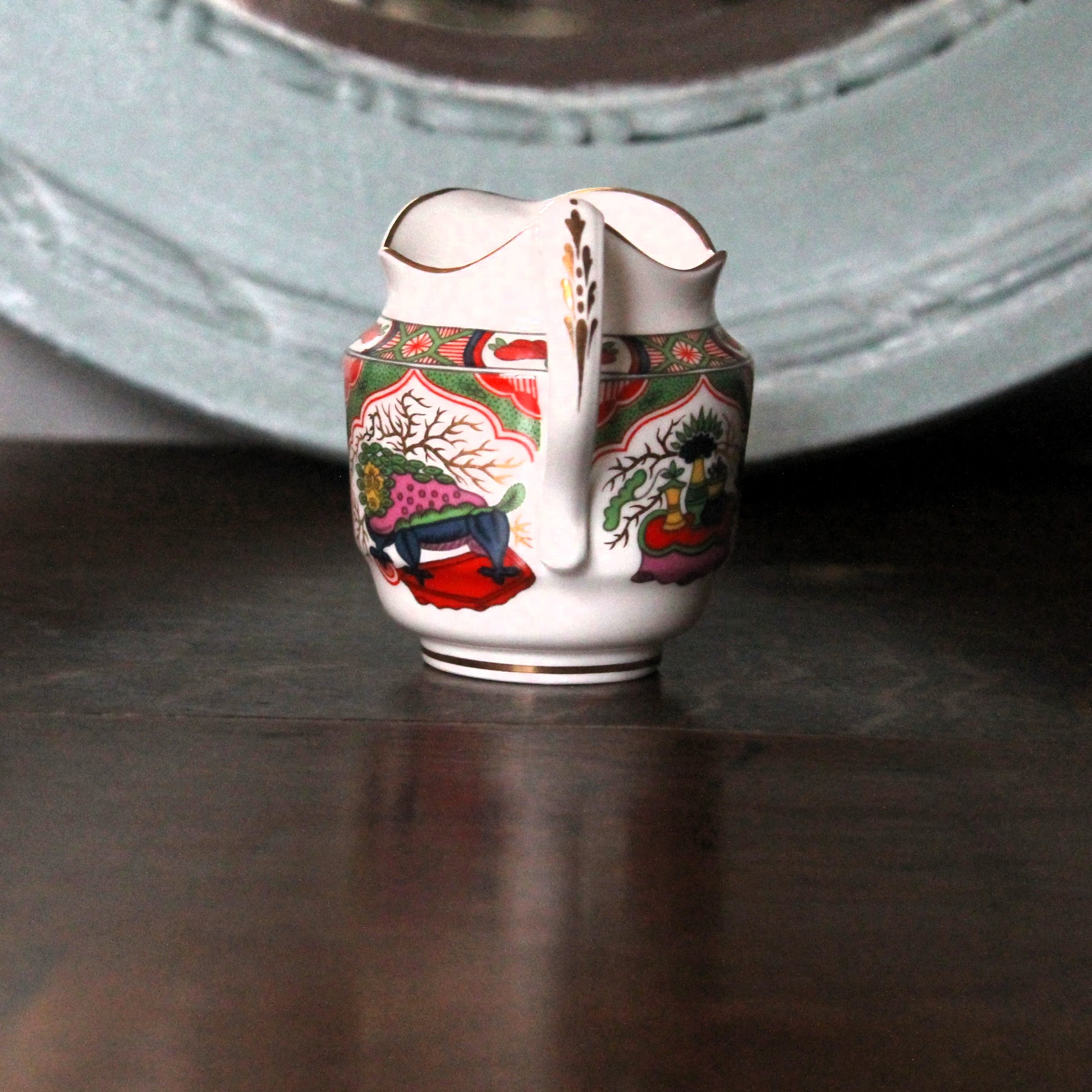 A small white porcelain creamer pitcher with Kilin pattern of floral and landscape illustration, featuring a gold-trimmed spout and handle, set on a dark wooden surface.