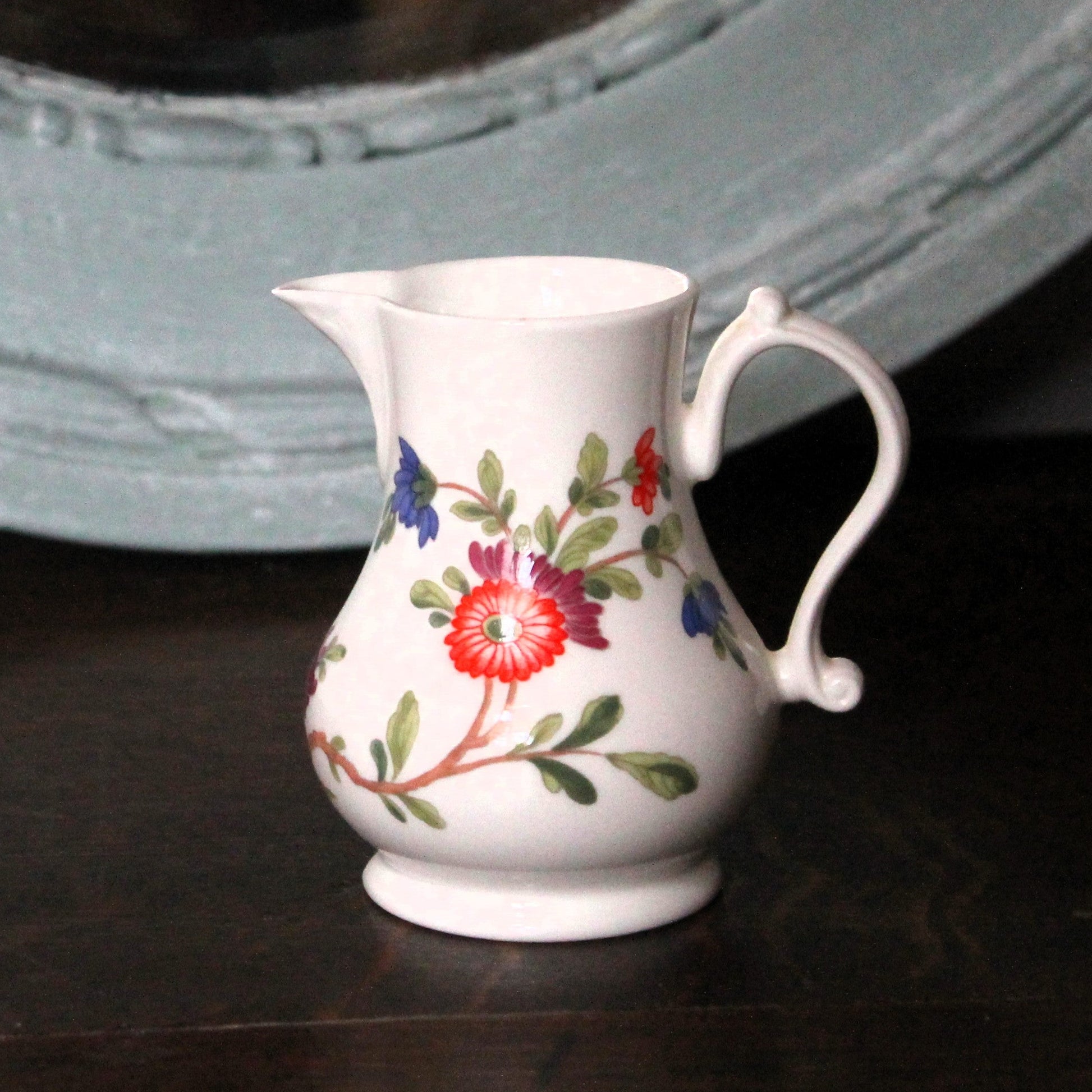 The Scratchcross jug,  small Royal Worcester white china jug with a floral pattern of red, blue, and green flowers and leaves, sitting on a dark wooden surface with a large, distressed light blue mirror in the background.