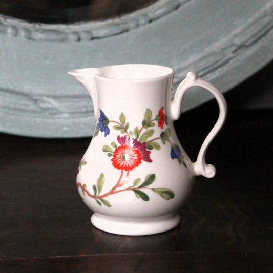 The Scratchcross jug,  small Royal Worcester white china jug with a floral pattern of red, blue, and green flowers and leaves, sitting on a dark wooden surface with a large, distressed light blue mirror in the background.