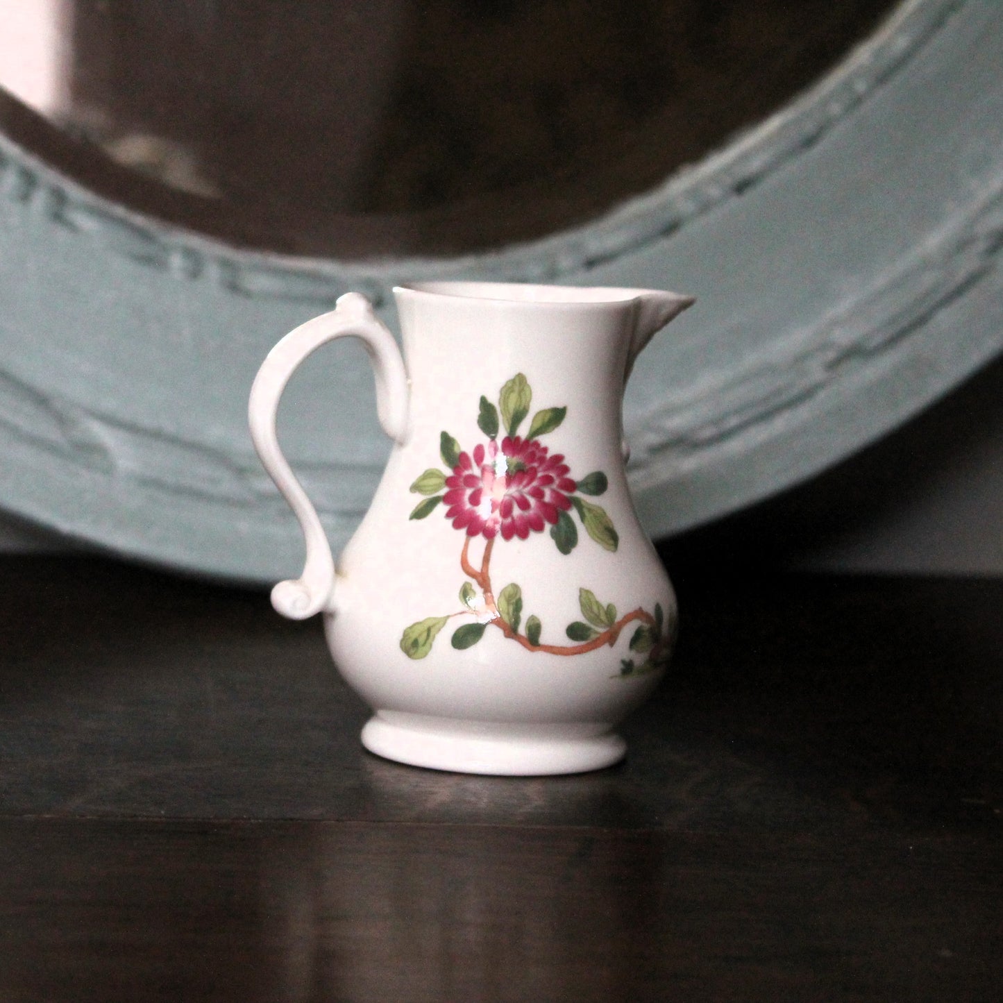 A small vintage Worceste porcelain creamer pitcher with a floral design of a pink and red flower with green leaves and a brown vine, set on a dark wooden surface with a blurred light blue mirror in the background.