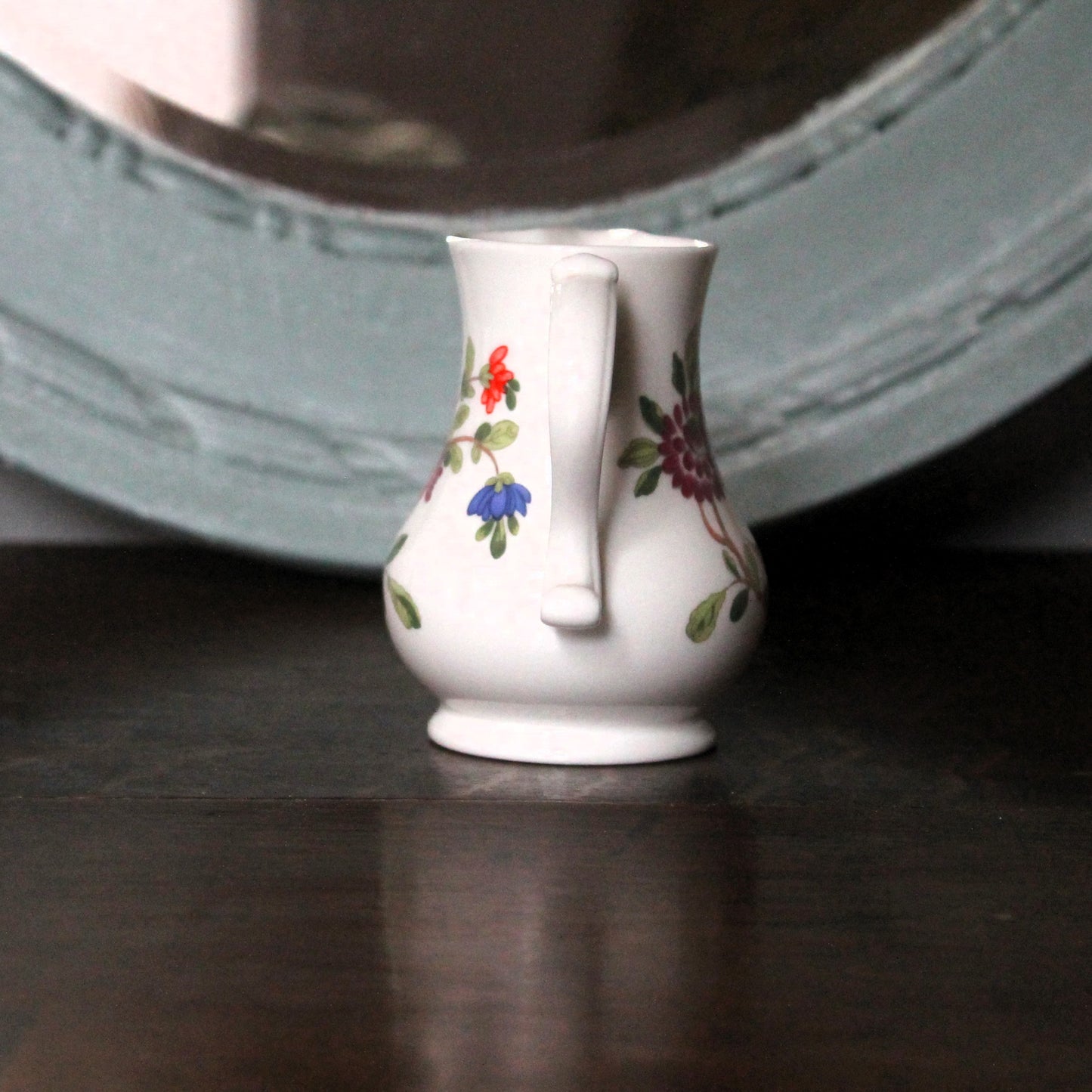 A small white porcelain creamer jug decorated with delicate floral patterns, standing on a dark wooden surface with a blurred mirror in the background.