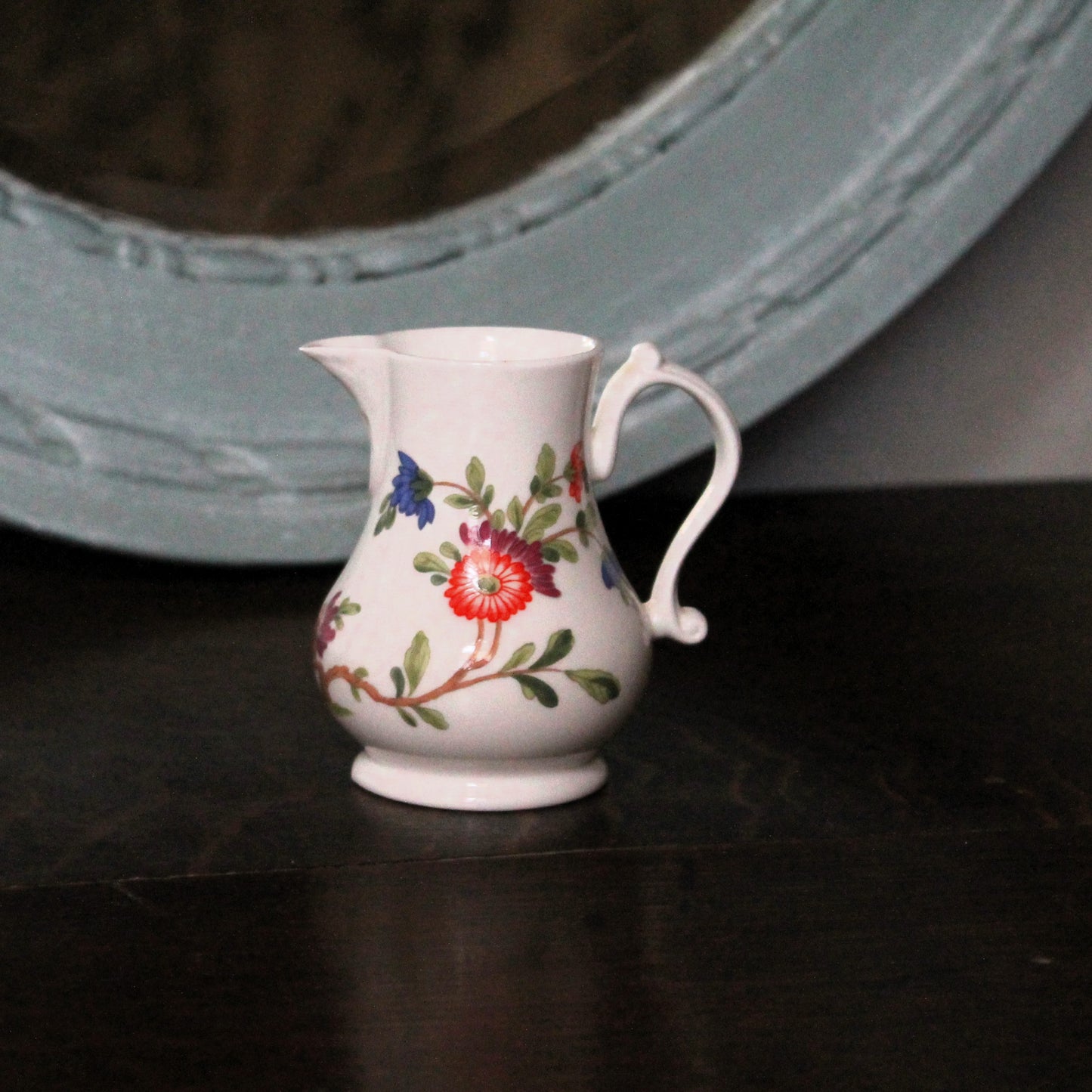 A small white porcelain creamer jug decorated with a colorful floral pattern, sitting on a dark wooden surface with a blurred light blue mirror in the background.