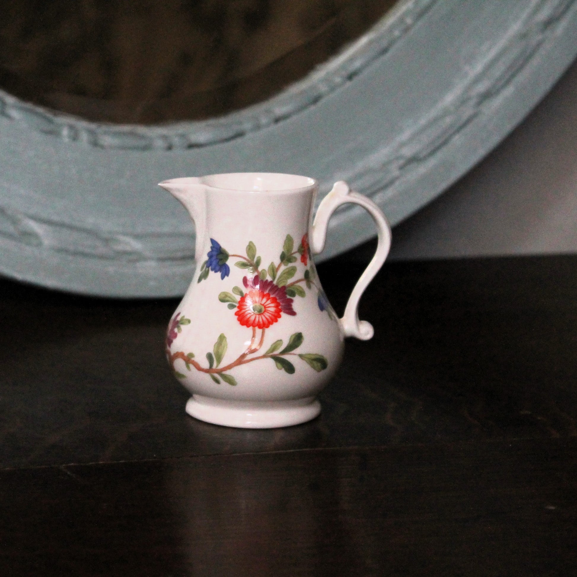 A small white porcelain creamer jug decorated with a colorful floral pattern, sitting on a dark wooden surface with a blurred light blue mirror in the background.