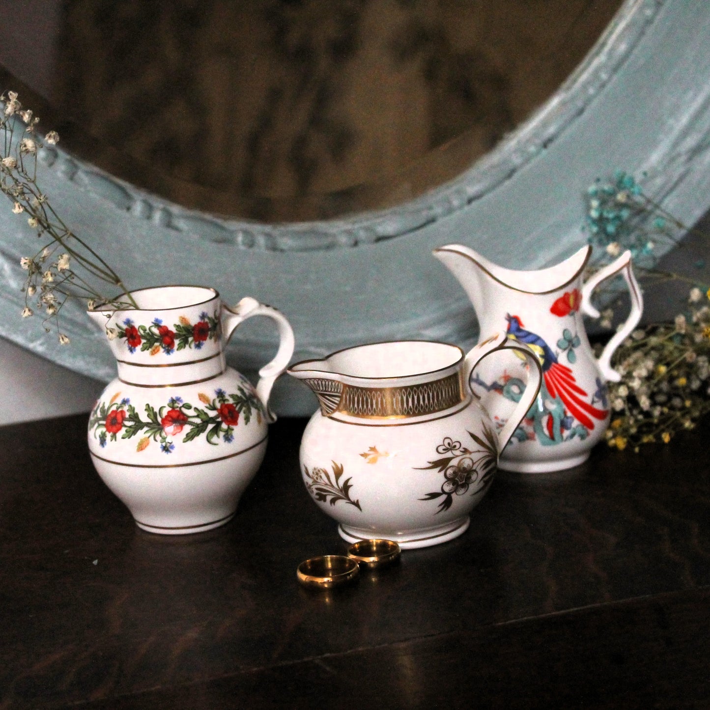 Three white porcelain pitchers with floral and bird motifs arranged on a dark tabletop in front of a large light blue mirror, with small dried flowers and gold rings as props.