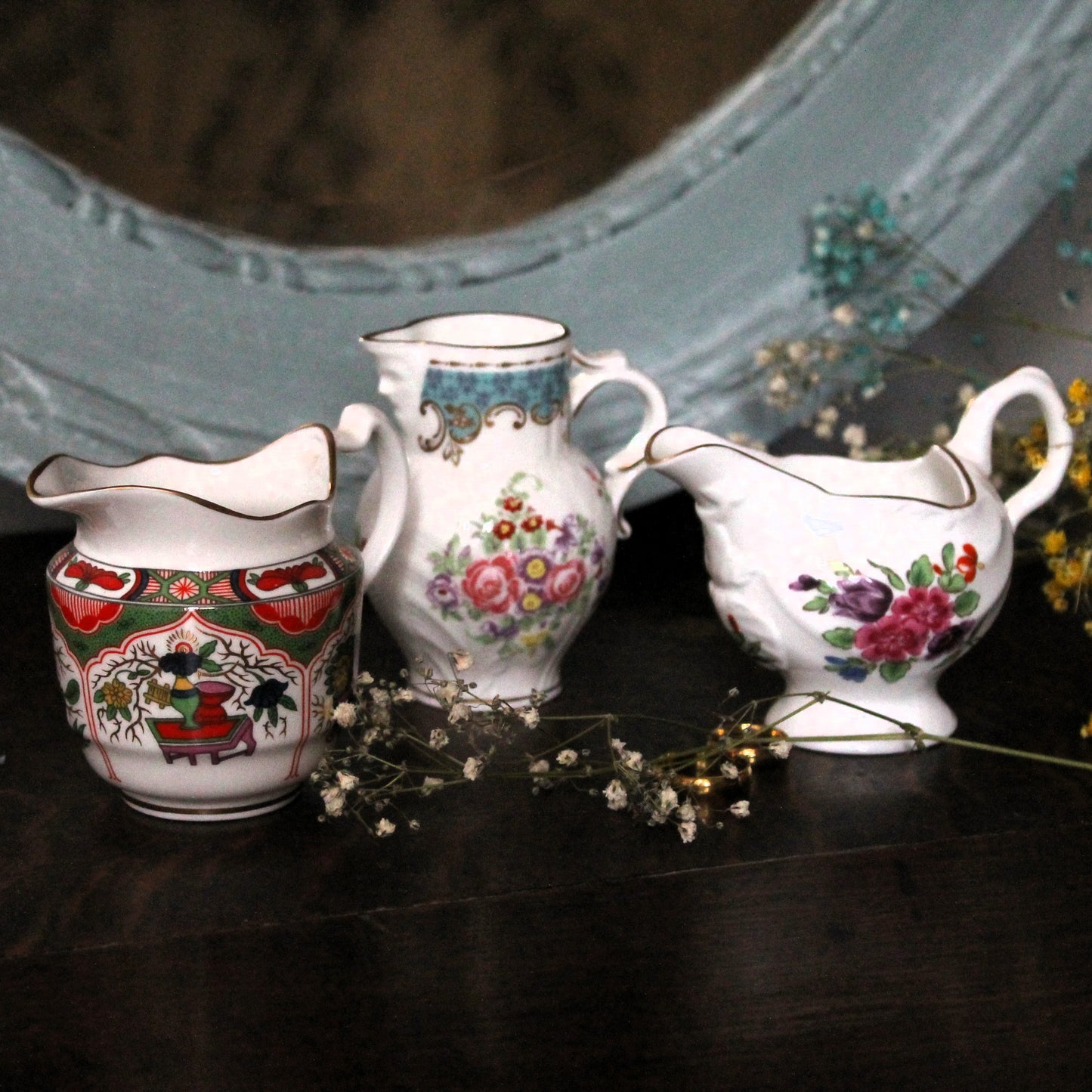 Three vintage white porcelain creamers jugs made by Compton & Woodhouse after Royal Worcester originals with colorful floral and hand-painted Chinese-style patterns, arranged on a dark wooden surface with dried flowers and a light blue mirror in the background.