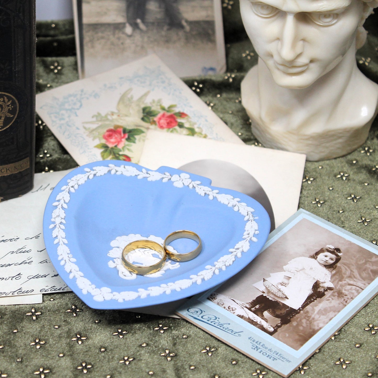 Blue heart-shaped vintage porcelain dish with gold rings on a table with vintage postcards and a bust.