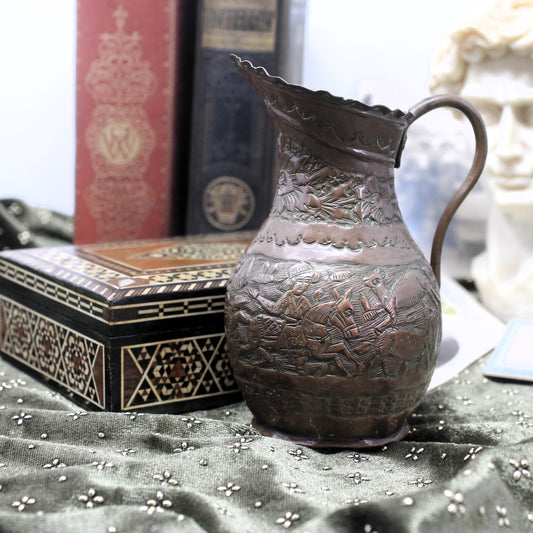 Small copper repousse jug with floral patterns and pastoral scene, set against a marqueterie box, on a brown cloth